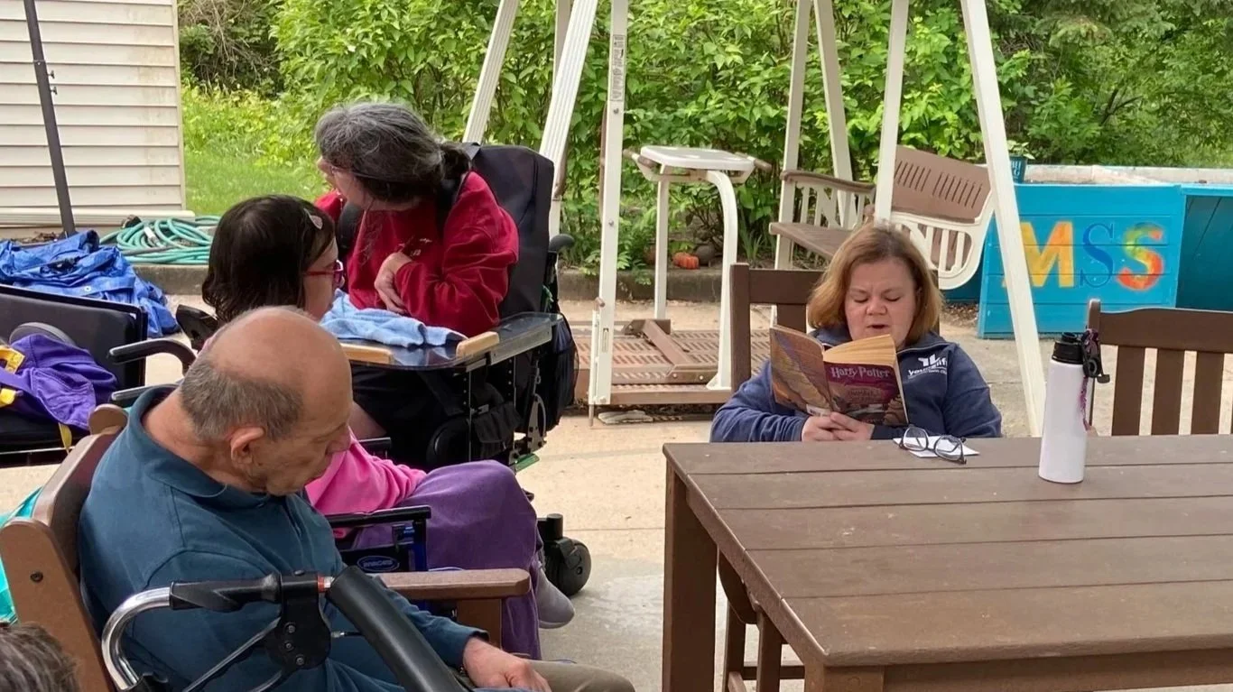 Jessica reading a Harry Potter book to an audience of three people in our programs, sitting around a patio table outside.