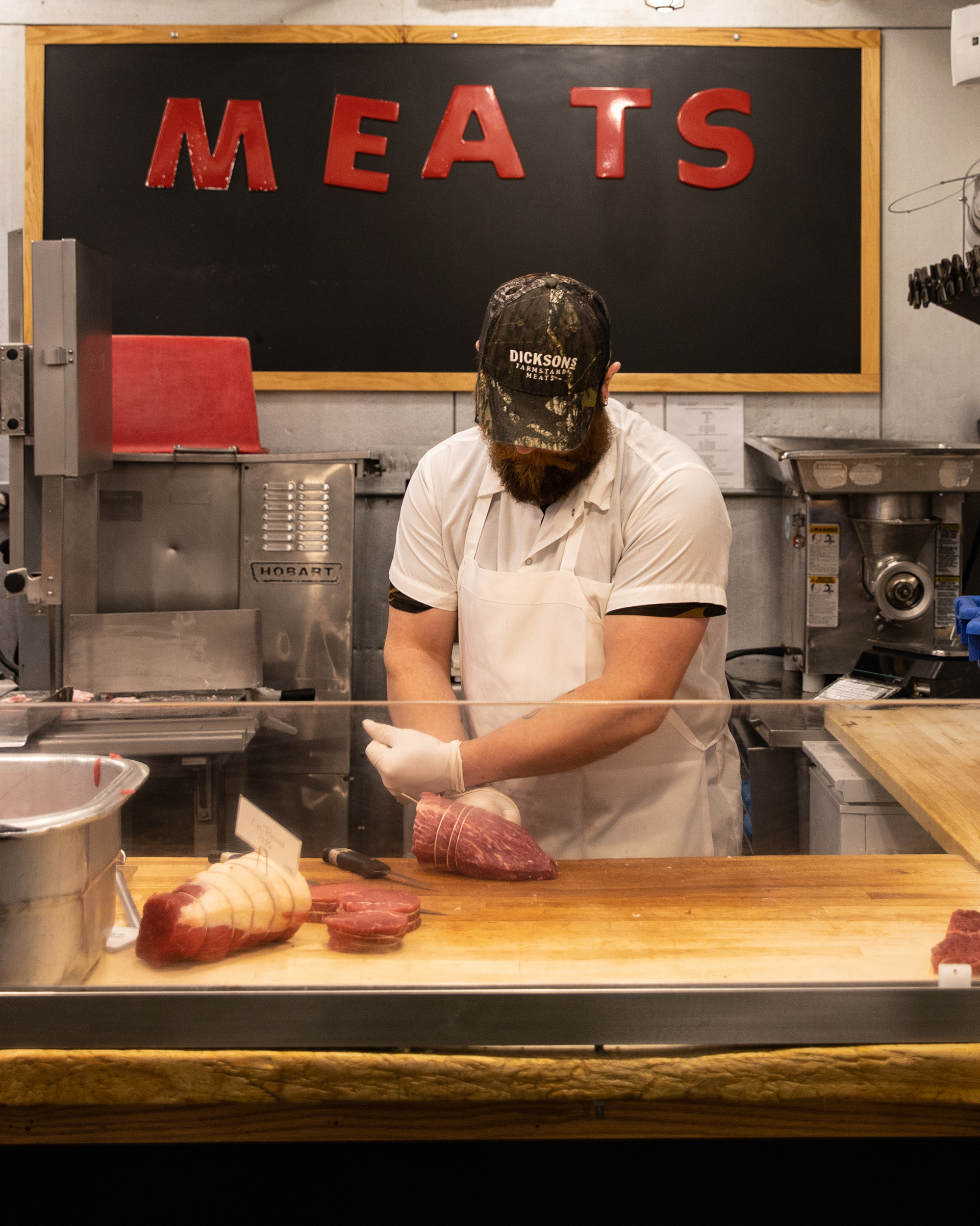 man standing in front of a chalk board with "meats" written in red on it carving cuts of meat 