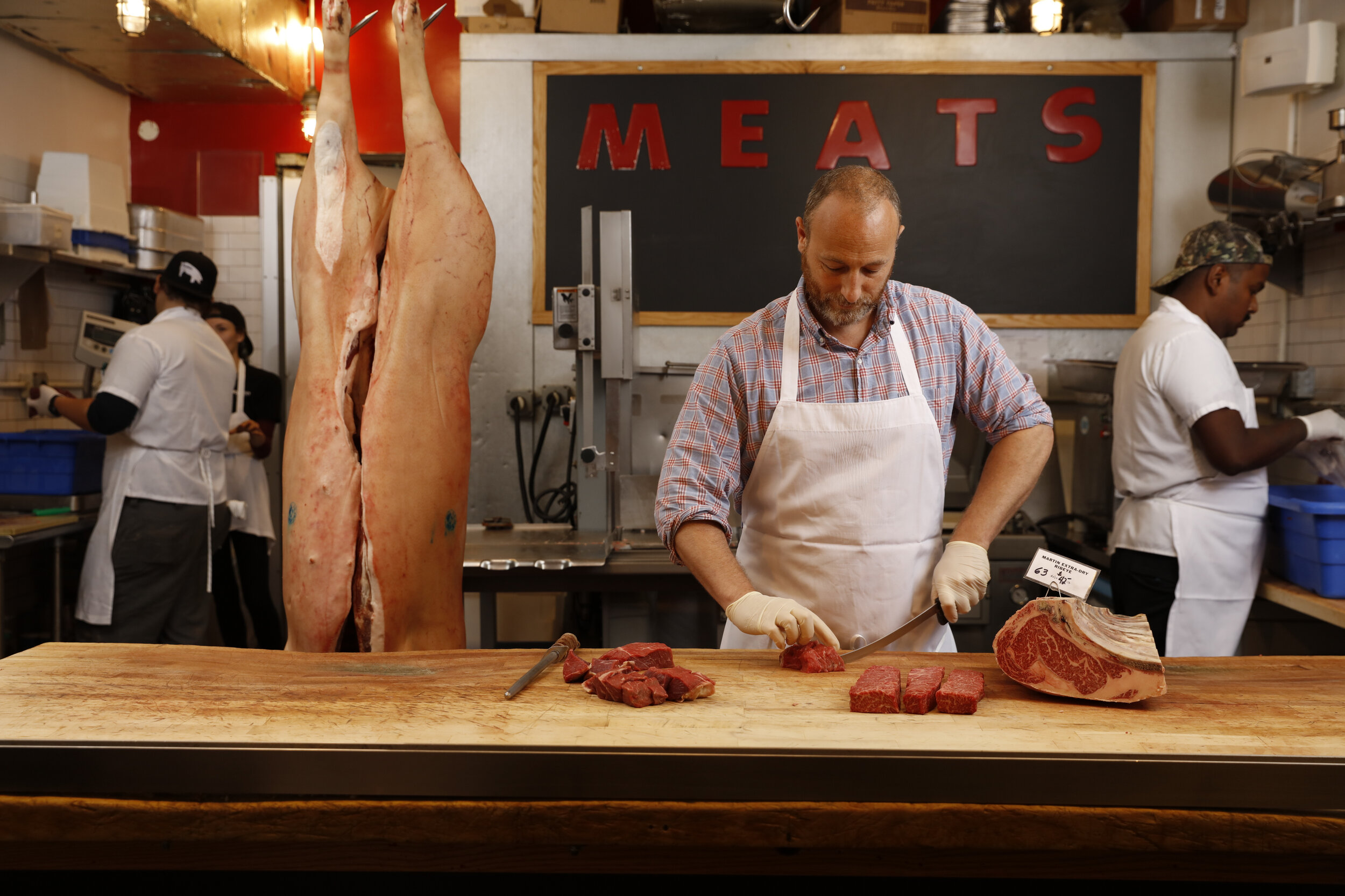 man in apron cutting meat in front of a black chalk board reading "meats"