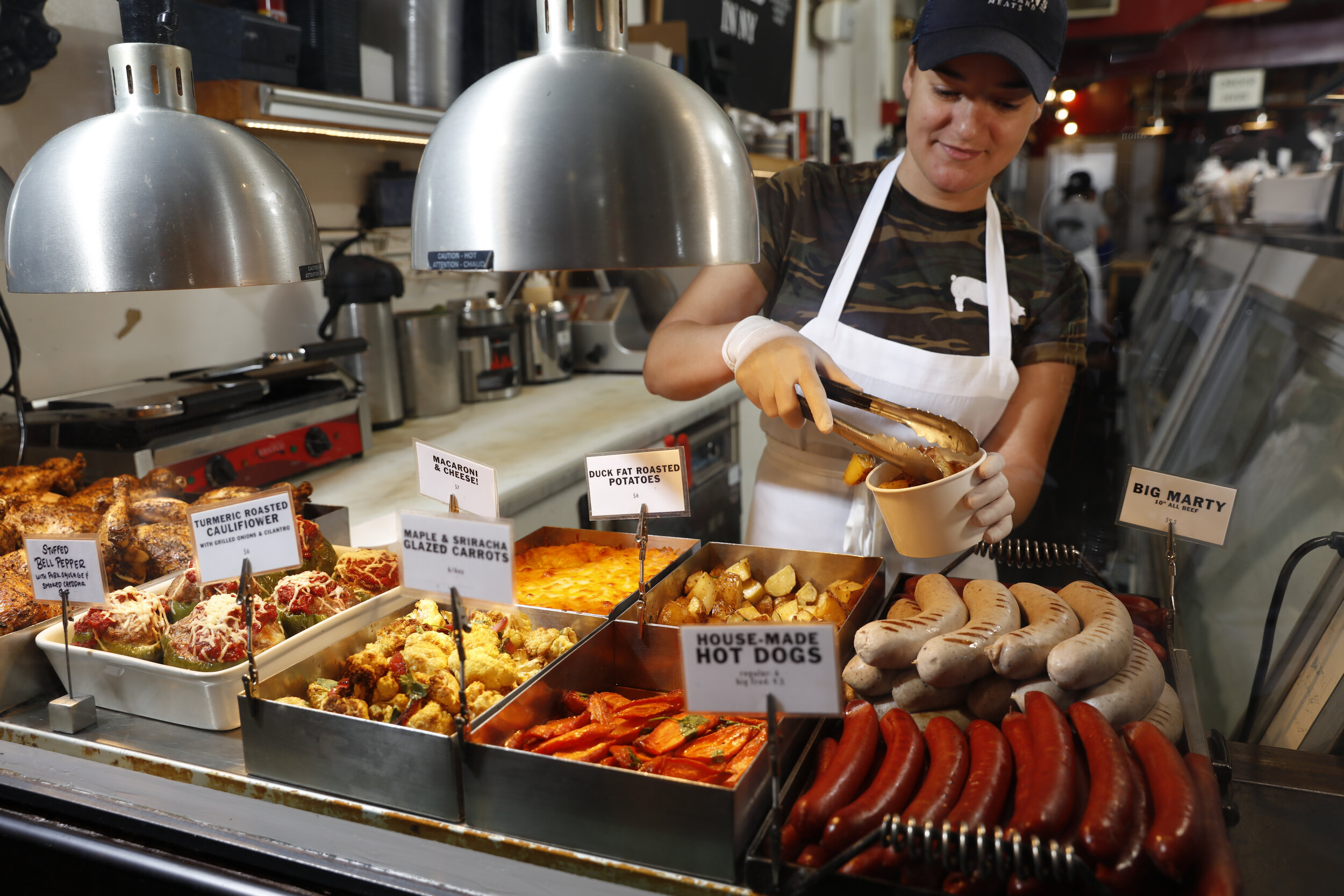 woman serving potatoes from a heat lamp with other prepared foods 