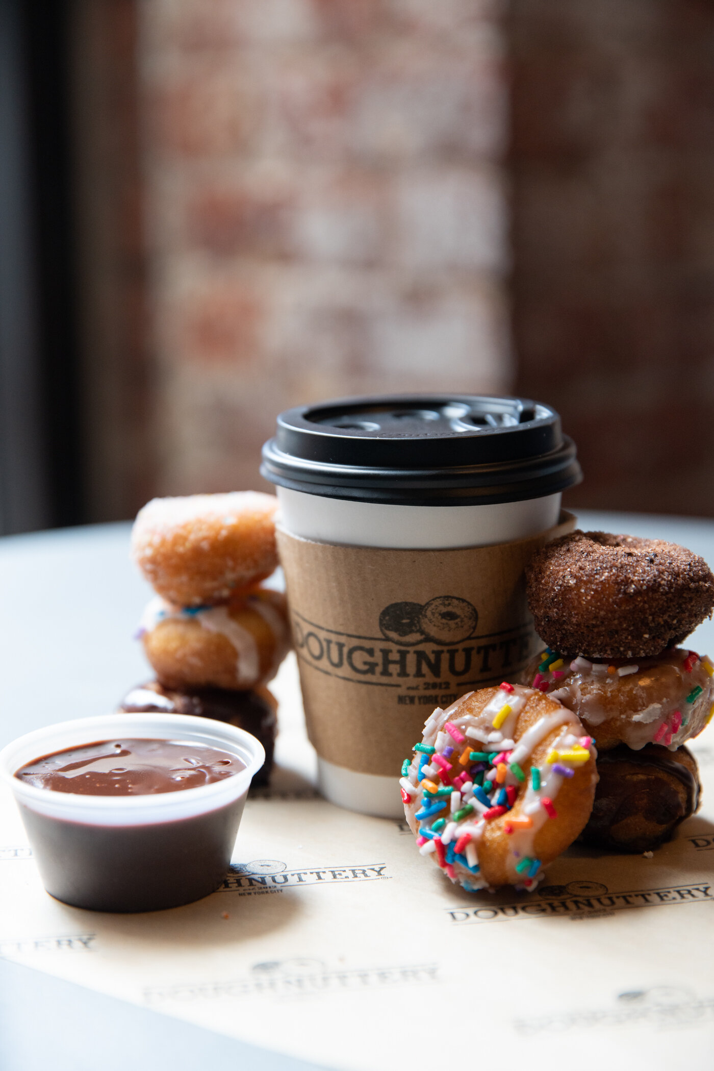 cup of coffee reading doughnuttery with mini dognuts surrounding it with a chocolate dipping sauce to the side 