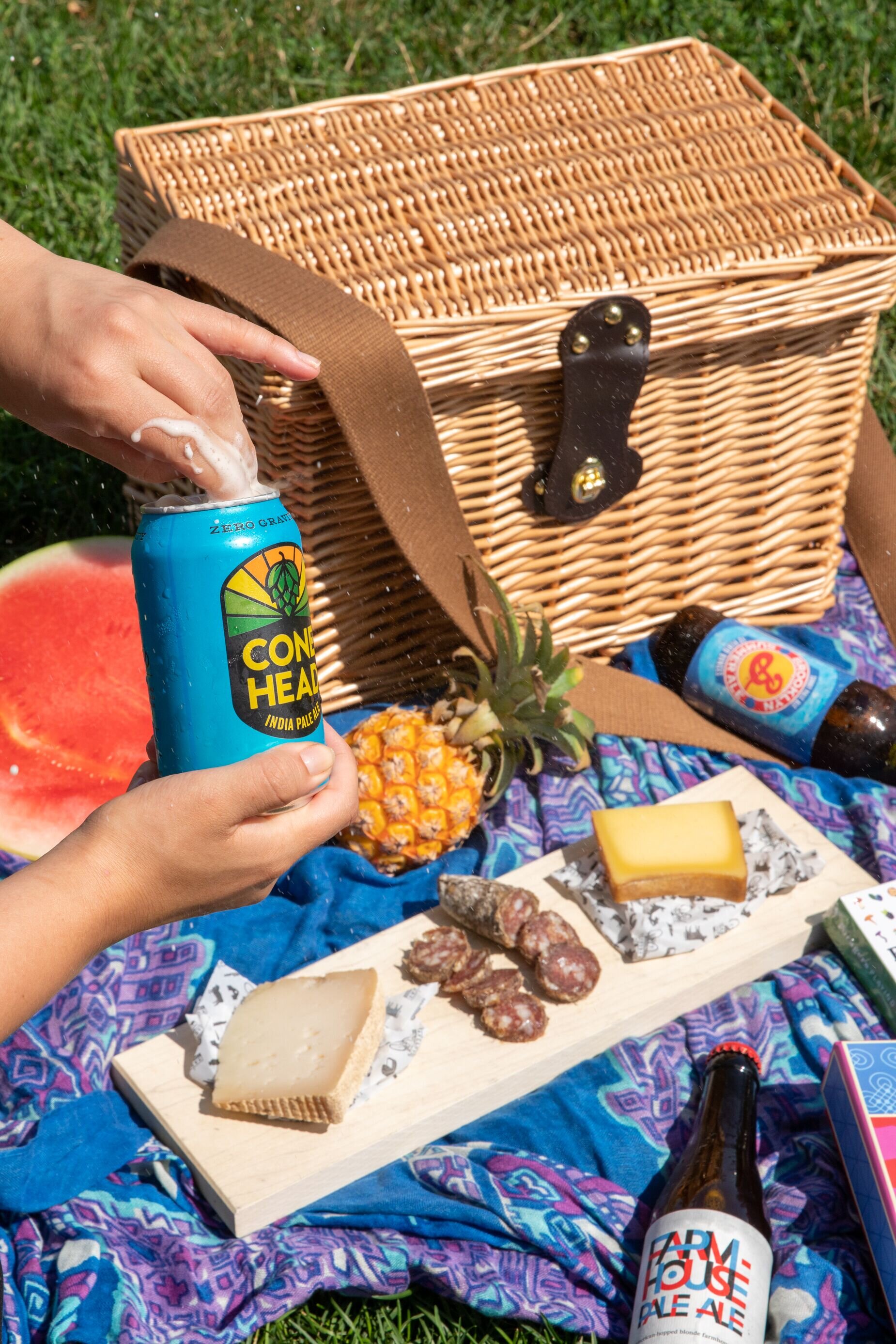 picnic basket on grass with a cheese board with meat and cheese and a hand opening a canned beer
