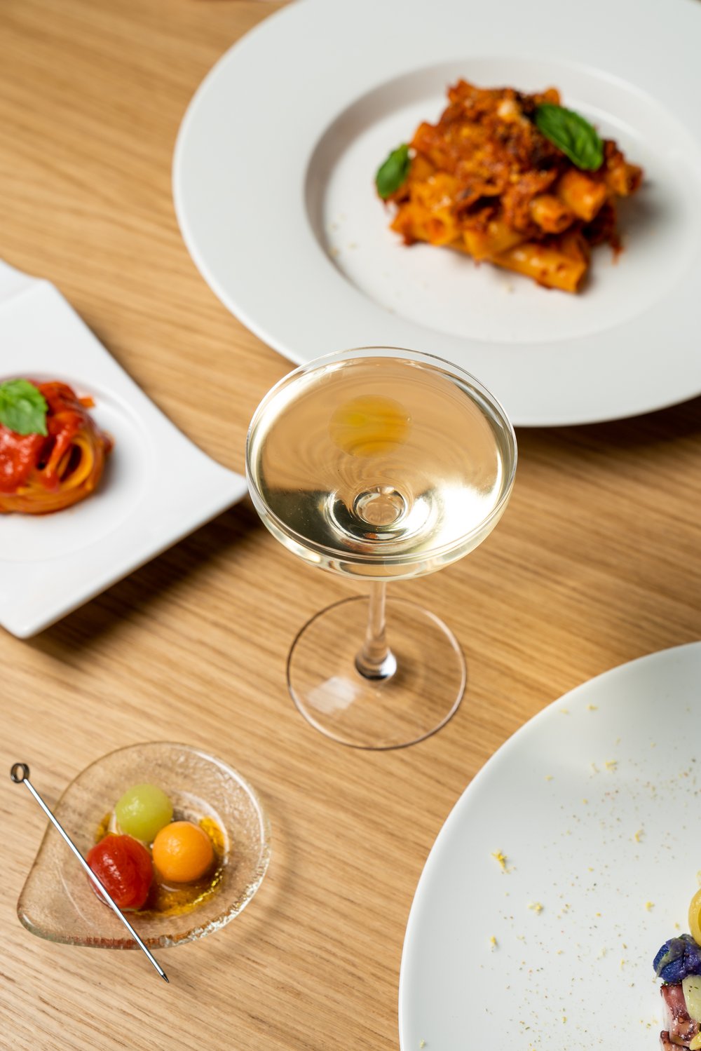 clear drink in cocktail glass on table with plates of pasta