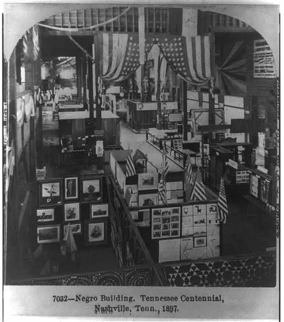 A black and white slide showing the inside of a fair exhibit building, with two American flags hanging from the ceiling. Exhibits of art and design can be seen on the ground, along with more flags.