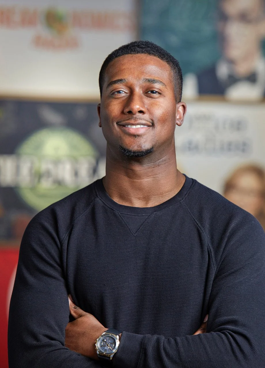 A smiling man with crossed arms wearing a black long-sleeve shirt and a wristwatch, standing indoors with a background of posters and pictures.