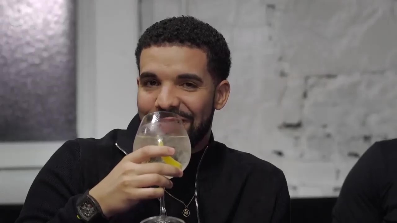 A man with curly dark hair and a beard is smiling while holding a large glass of what appears to be a cocktail or beverage with lemon slices inside.