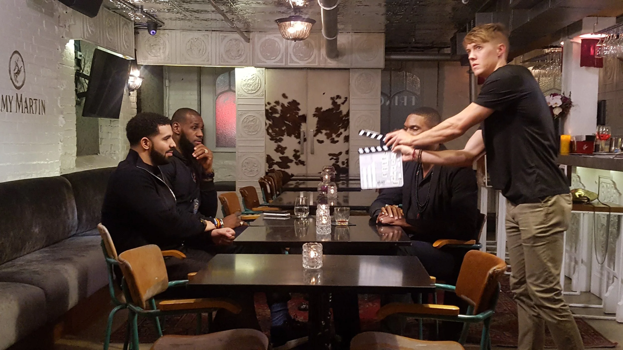Four men are sitting at a dark table in a restaurant or bar, with a waiter or crew member standing and holding a clapperboard, possibly filming a scene. The setting has dim lighting, decorative wall panels, and a relaxed atmosphere.