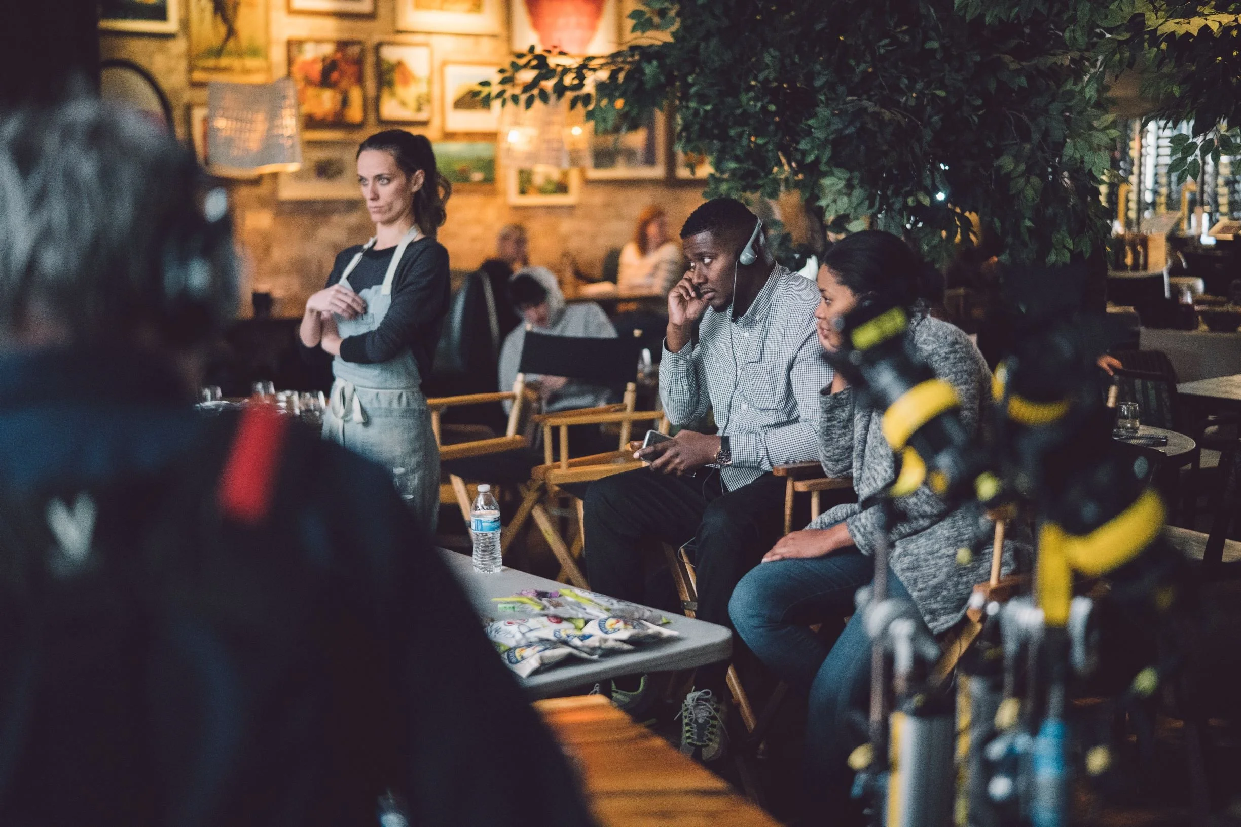 A woman waitress in apron stands near a table in a busy restaurant, observing customers. Two diners, a man wearing headphones and a woman, are seated at a table, with the man looking at his phone. The restaurant has a warm ambiance with artwork on the walls and greenery, with other customers visible in the background.