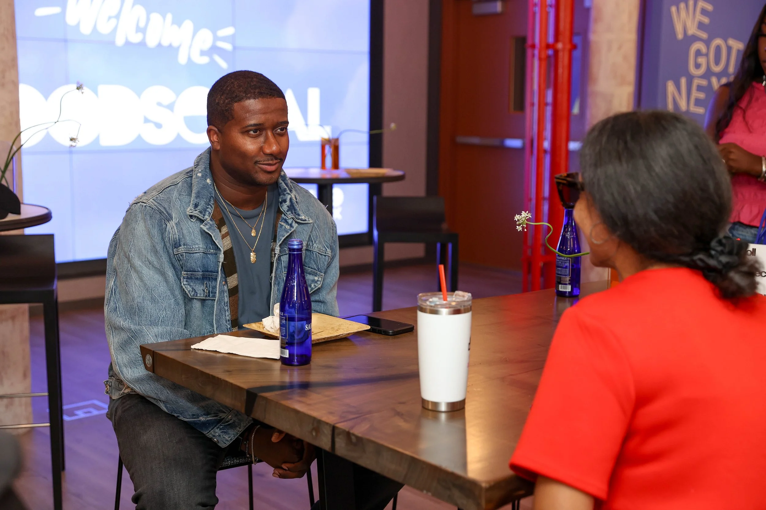 A young man and an older woman sitting at a restaurant table, engaged in conversation. The young man is wearing a denim jacket and has a drink and a bottle of water in front of him. The woman is dressed in a red top, and there are flowers in vases on the table.