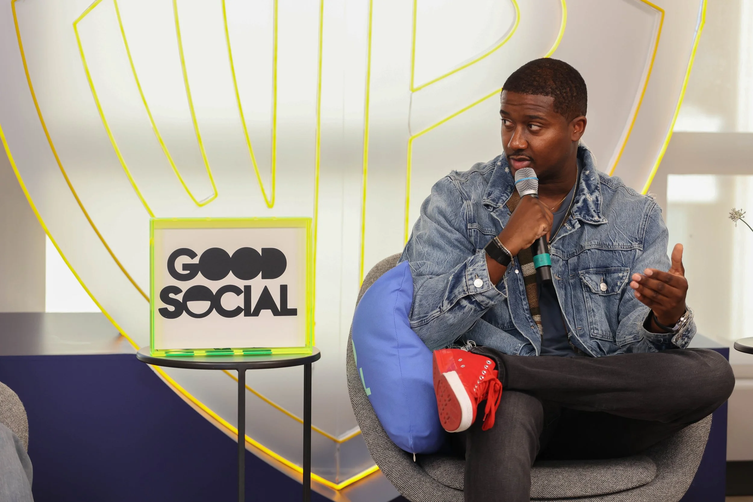 Man sitting in a gray chair holding a microphone, wearing a denim jacket, black pants, and bright red sneakers with white soles, in a modern room with a neon yellow heart outline in the background and a sign that says "Good Social" on a small side table.