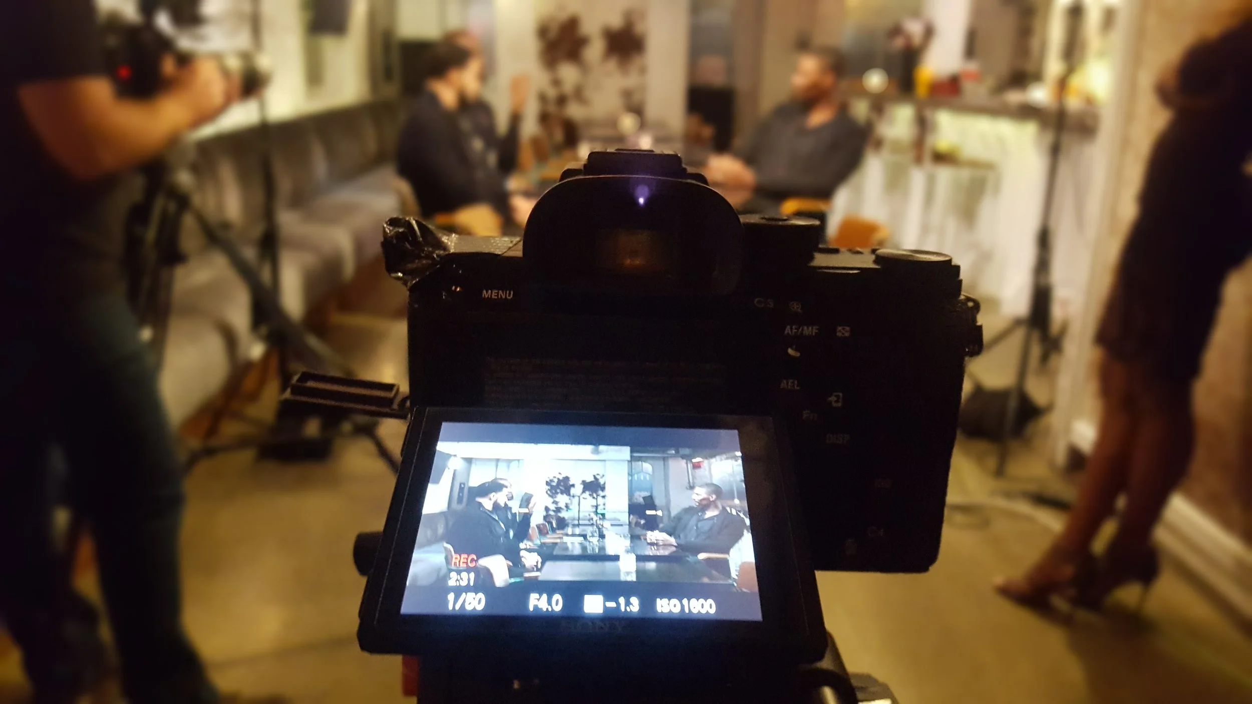 Camera capturing a scene with two people in a discussion, with a crew member in the background, and the camera's screen showing the recorded scene.