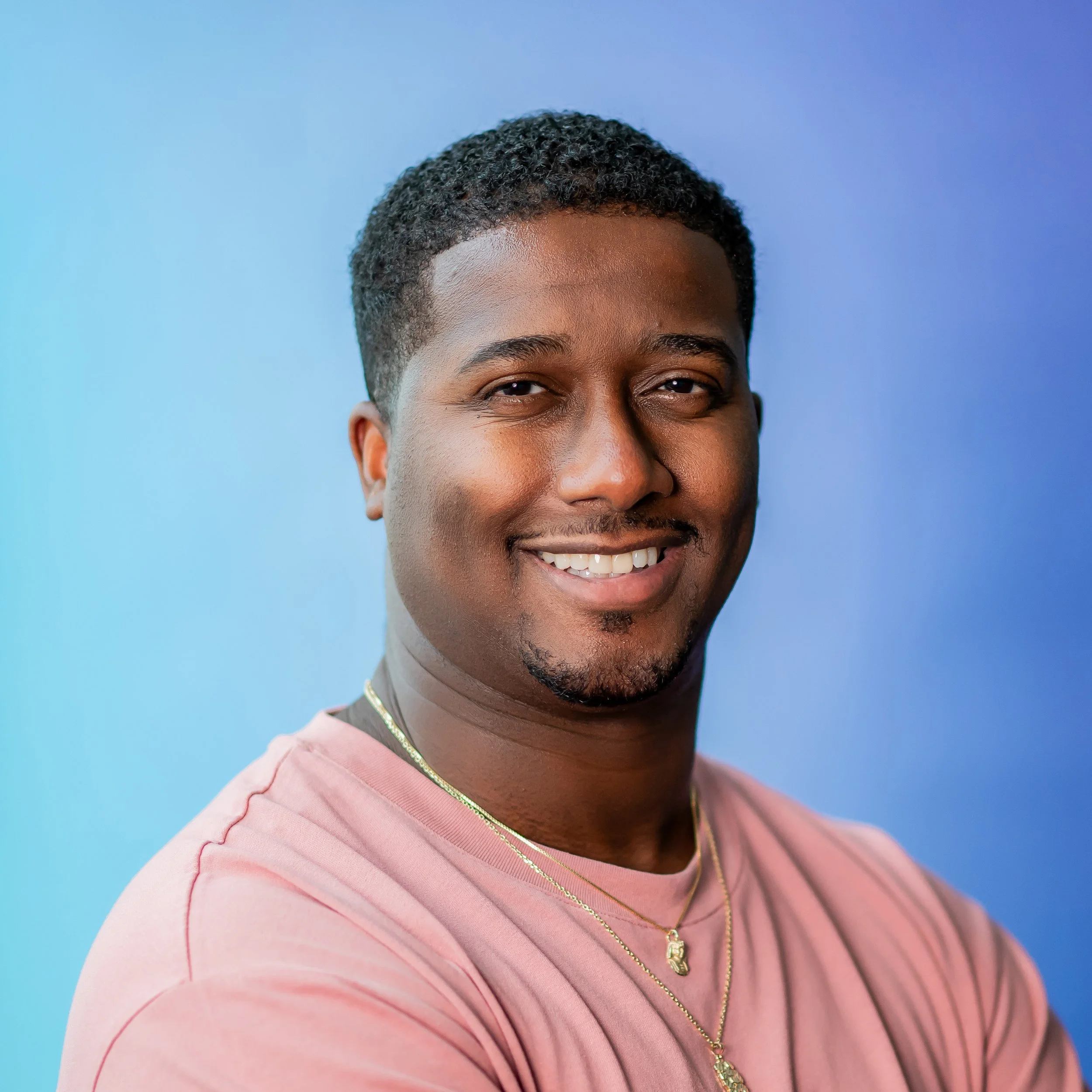 A smiling man with short curly black hair, wearing a pink shirt and gold necklaces against a blue background.