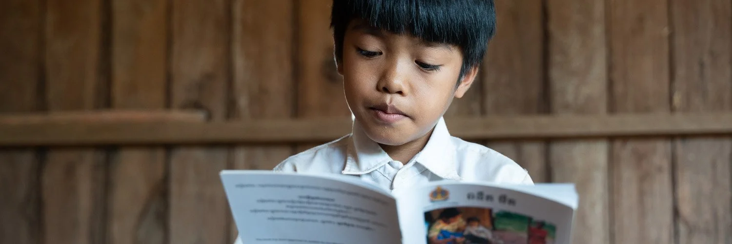 A young boy with black hair wearing a white shirt, reading a brochure or booklet in a wooden room.