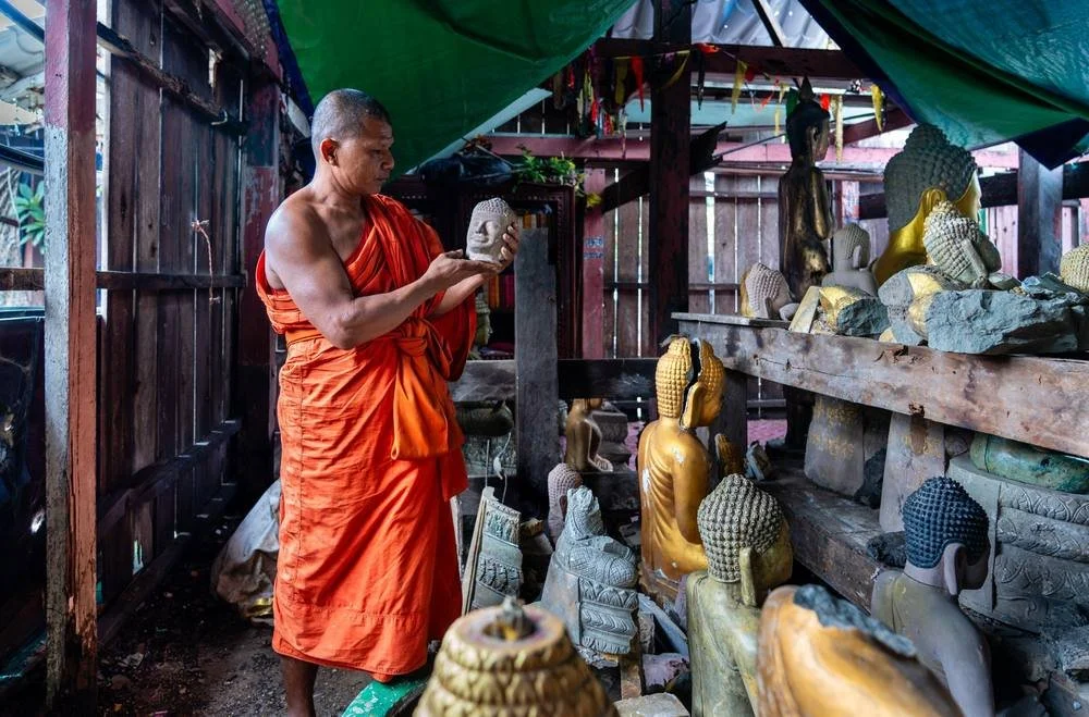 Abbot Sun Sang holding a broken stone Buddha head among damaged statues at Wat Keo Sekha Kiri Svarak, Preah Vihear