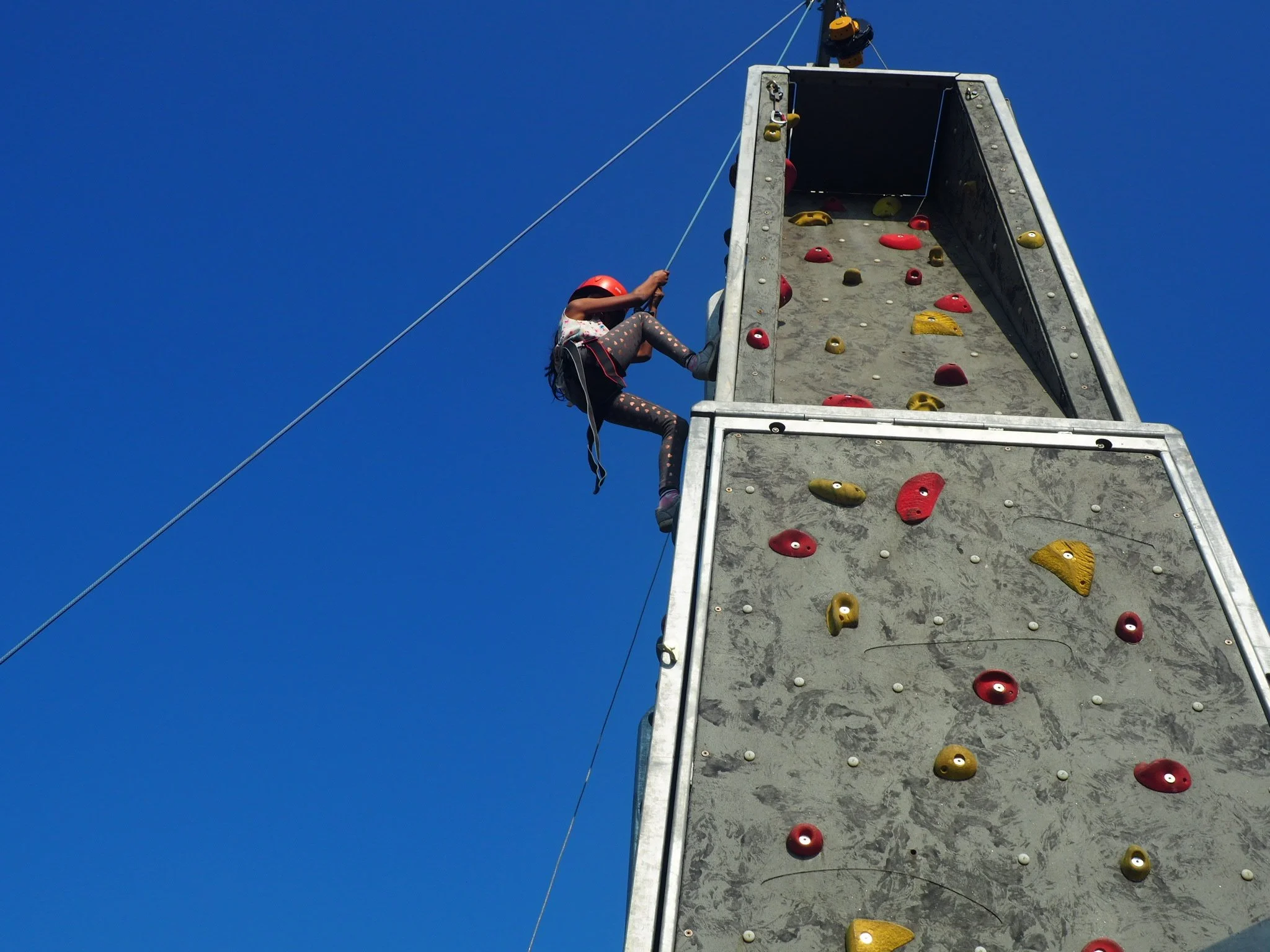 A child reaching the top of a climbing wall, silhouetted against a blue sky