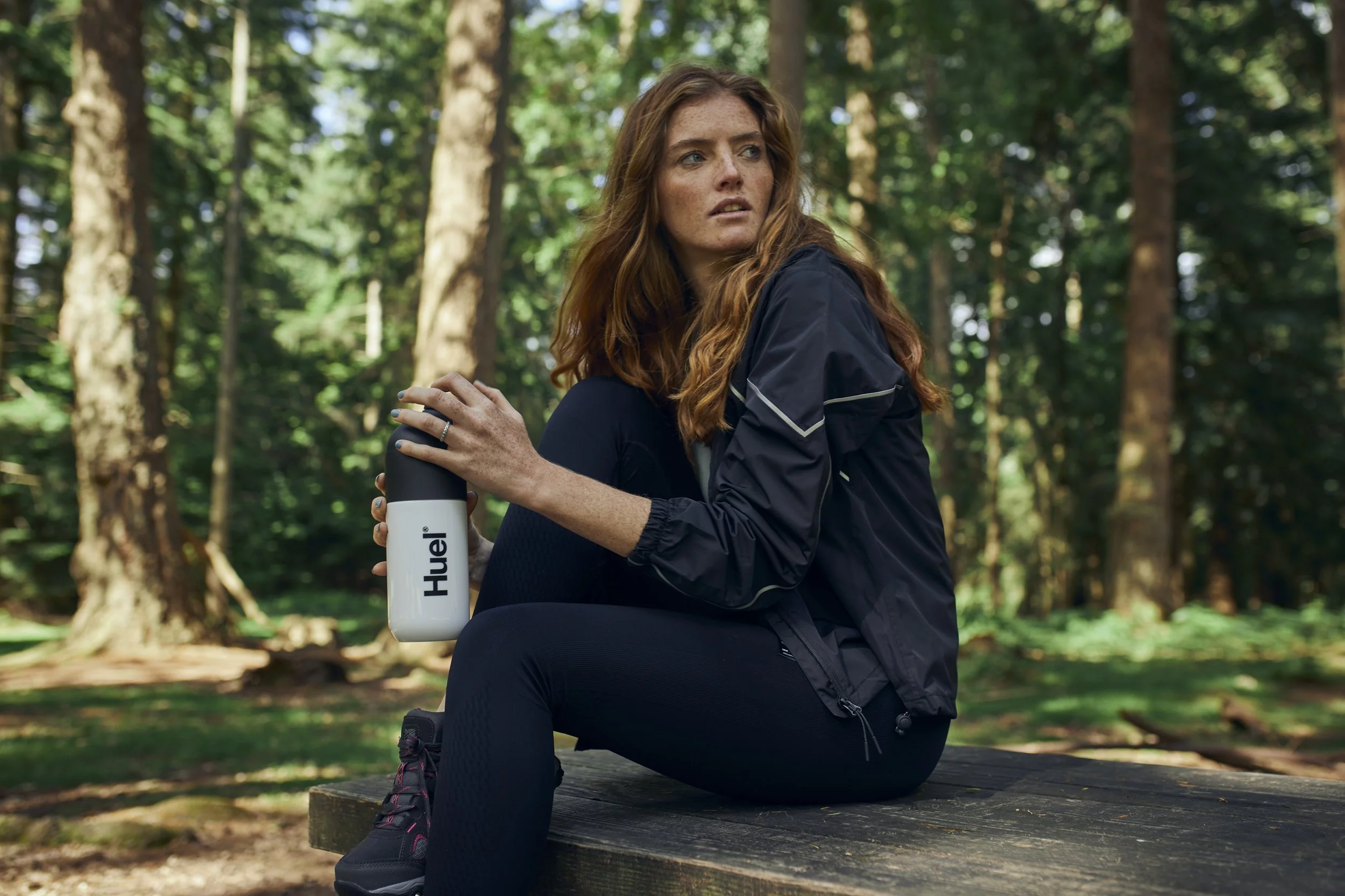 A women sits a a woods, surrounded by large tall trees. She drinks from a bottle of Huel. Lifestyle Photography by Jon Enoch