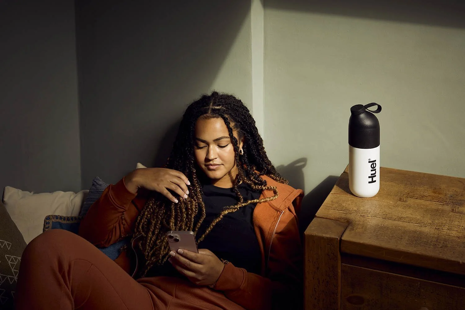 A young women sit on her bed using her phone, a bottle of huel next to her in this lifestyle image by Jon Enoch