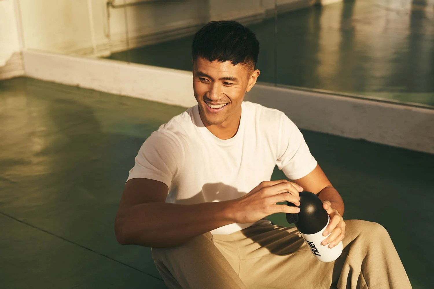 A man unscrews a bottle of Huel which he sits on the floor of a gym. A shaft of warm light hitting him in this advertising photo by Jon Enoch