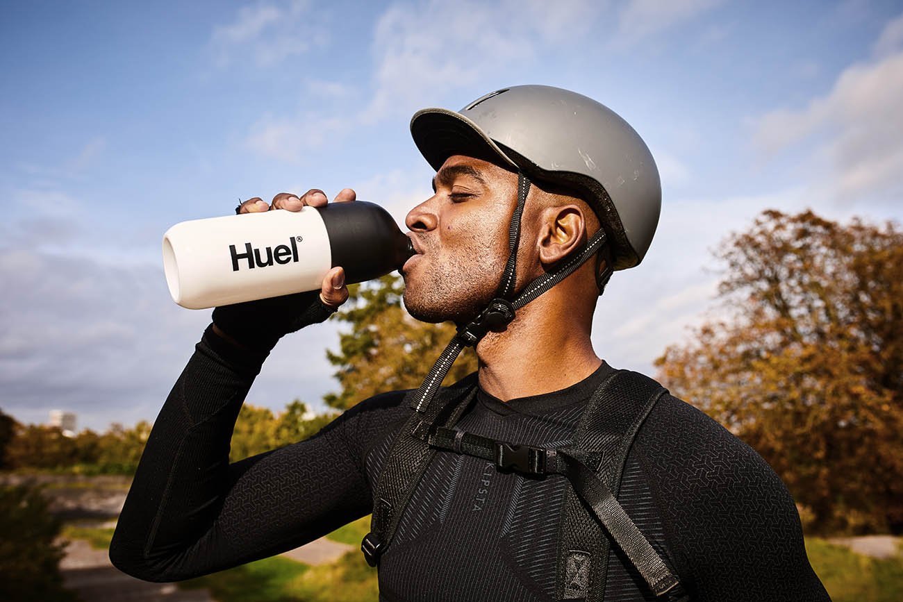 A man wearing a bike helmet drinks from a Huel Shaker in this lifestyle image by Jon Enoch
