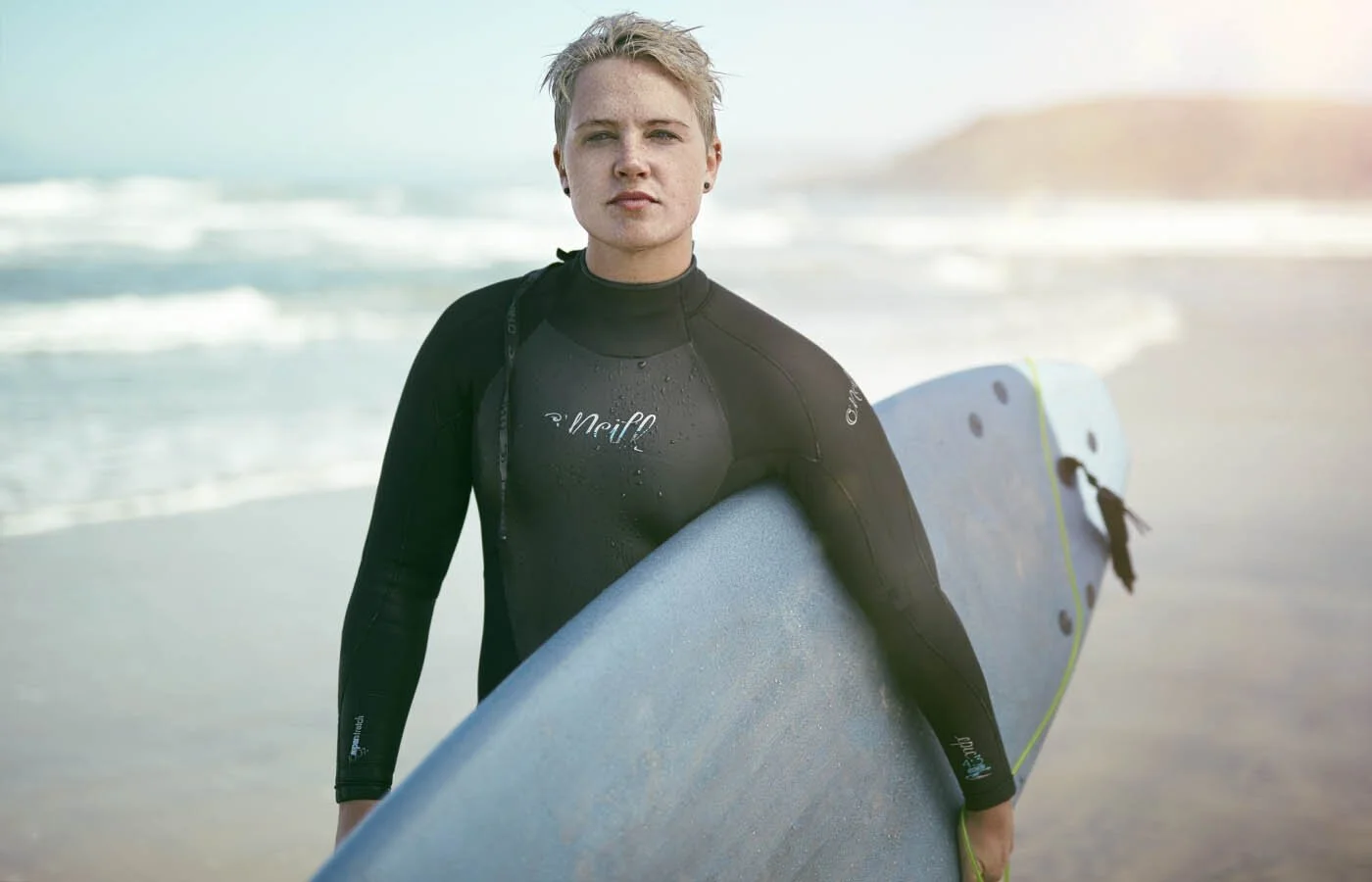 A women holds her surf board after a surf on a remote beach in California as the sun rises over the beach. Lifestyle photography by Jon Enoch.