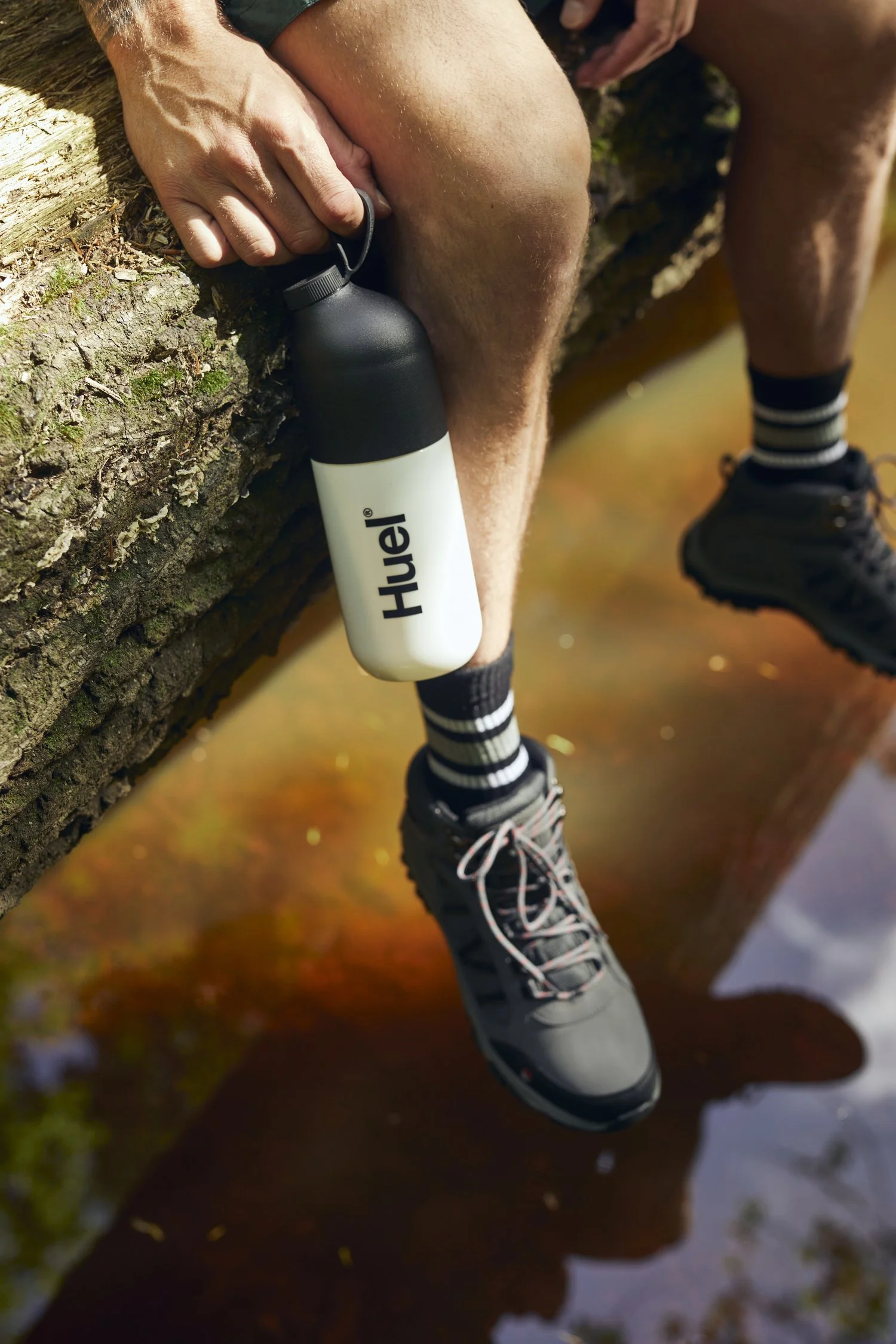 A mans legs dangle off a fallen tree over a river. A bottle of Fuel in hand. Images by lifestyle photographer Jon Enoch.