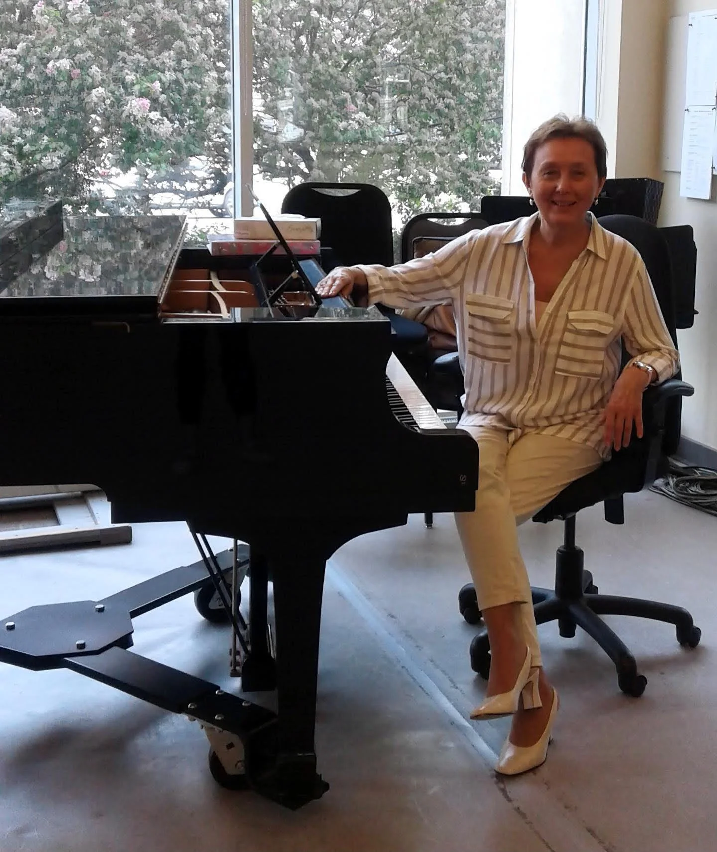 A woman in a striped shirt and white pants sitting on a black office chair next to a grand piano in a room with large windows and trees outside.