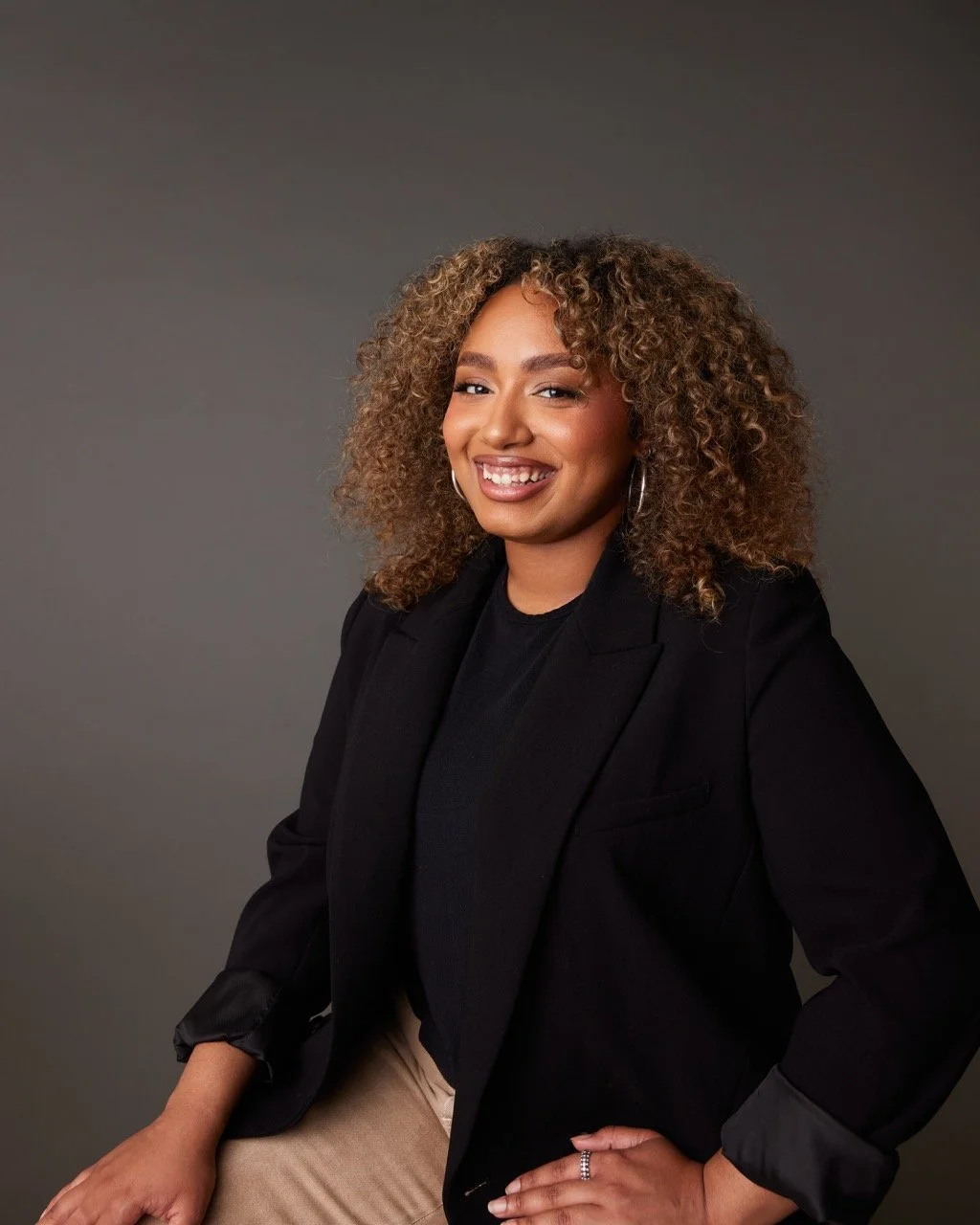 A smiling woman with curly hair, wearing a black blazer and beige pants, posed with one hand on her hip against a neutral grey background.