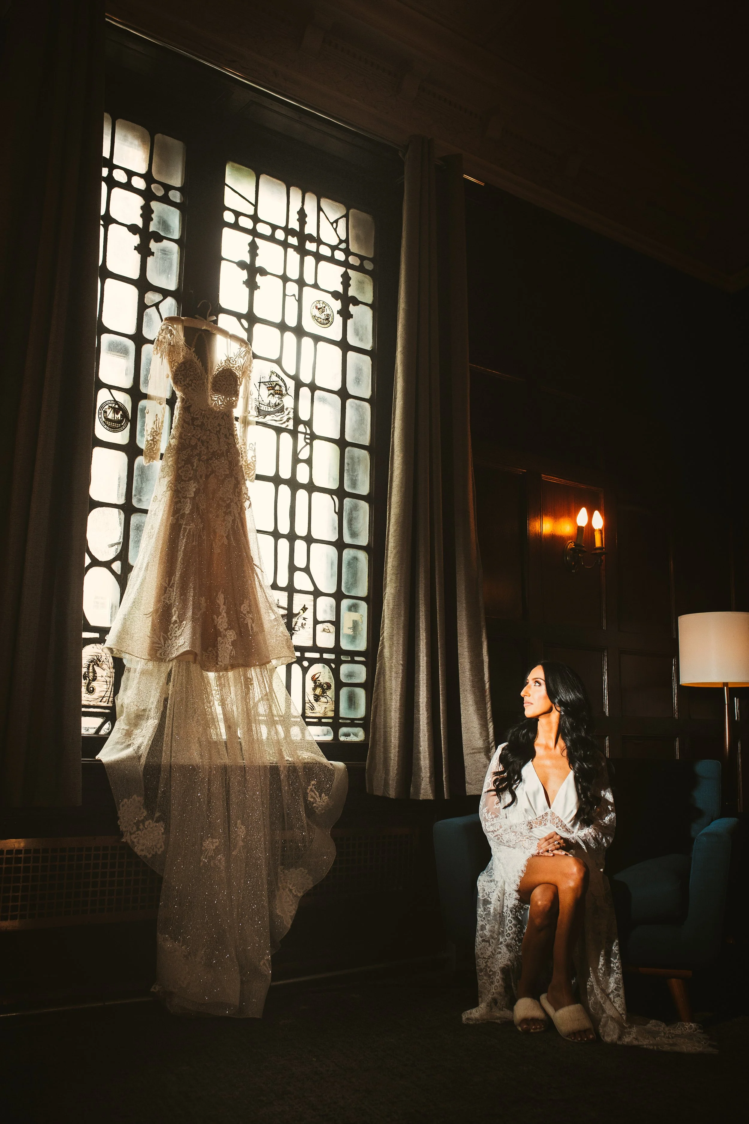 Bride sitting and looking up at her wedding dress hanging from the tall window