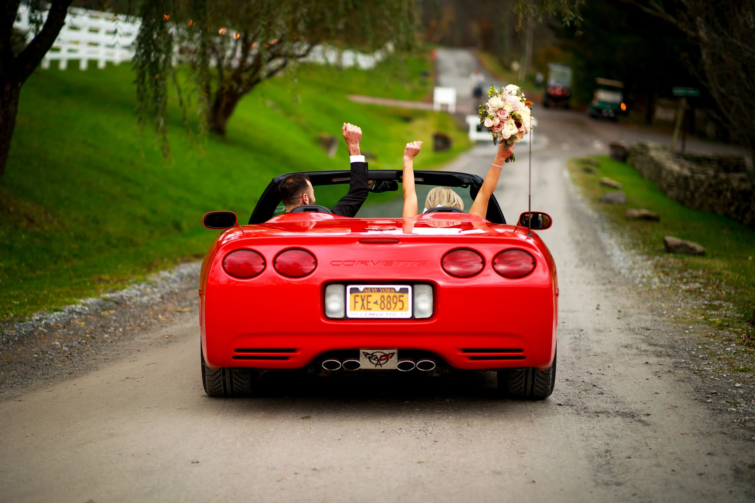 Bride and groom cheering with their hands up as they drive away after the ceremony in a red sports car