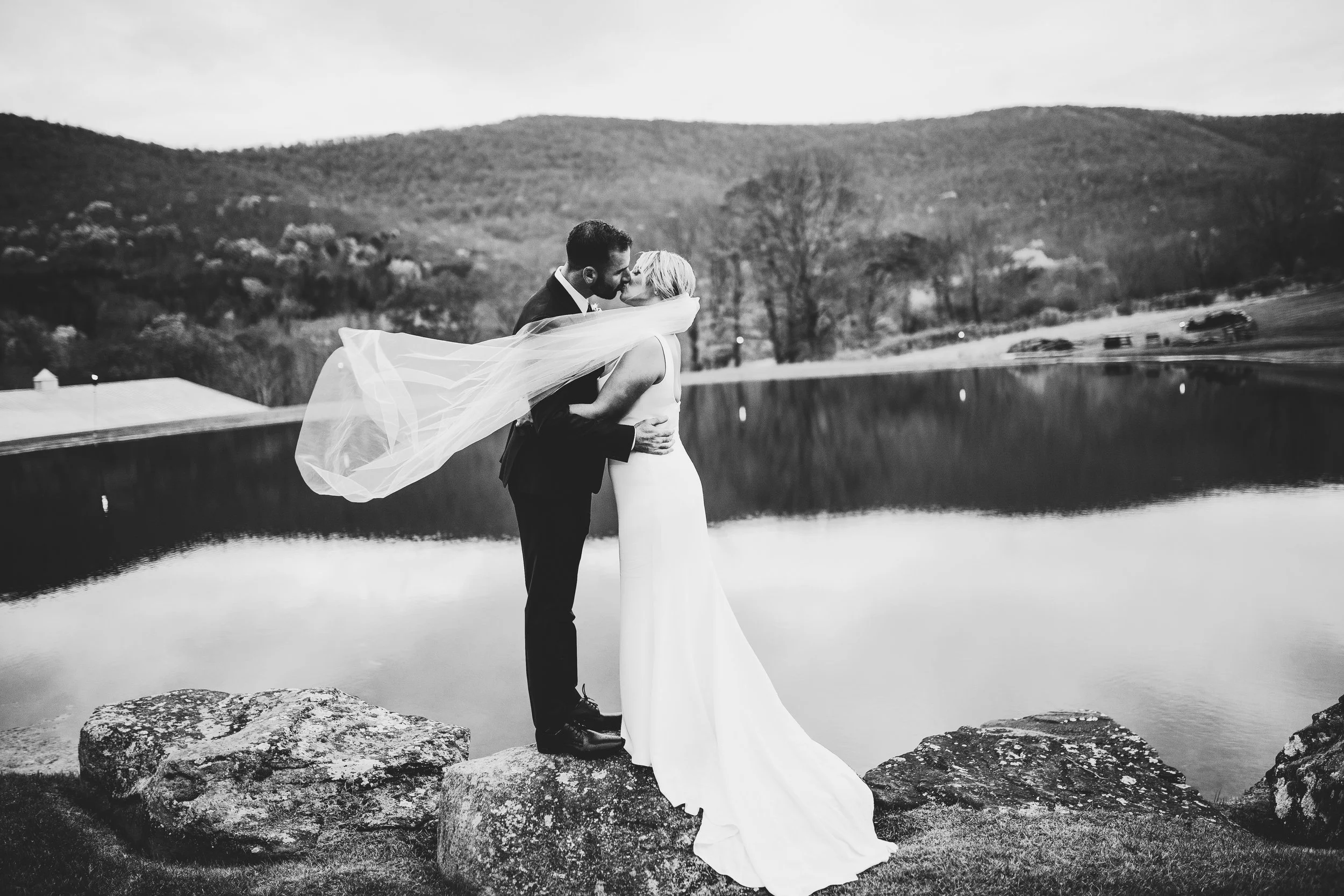 Bride and groom kissing in front of a pond with the bride's veil blowing in the wind
