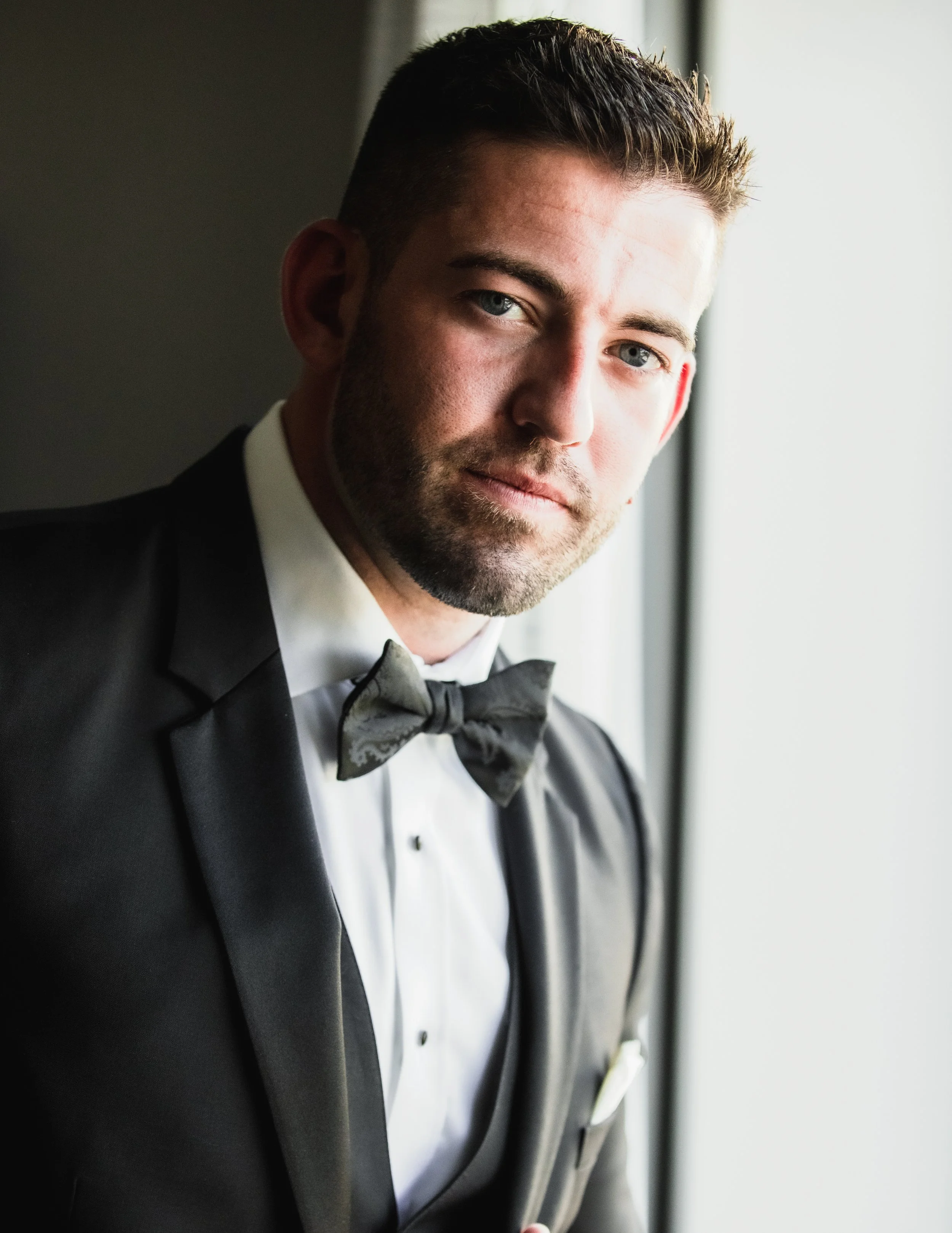 Groom looking at the camera while standing in a window, while wearing his black tux