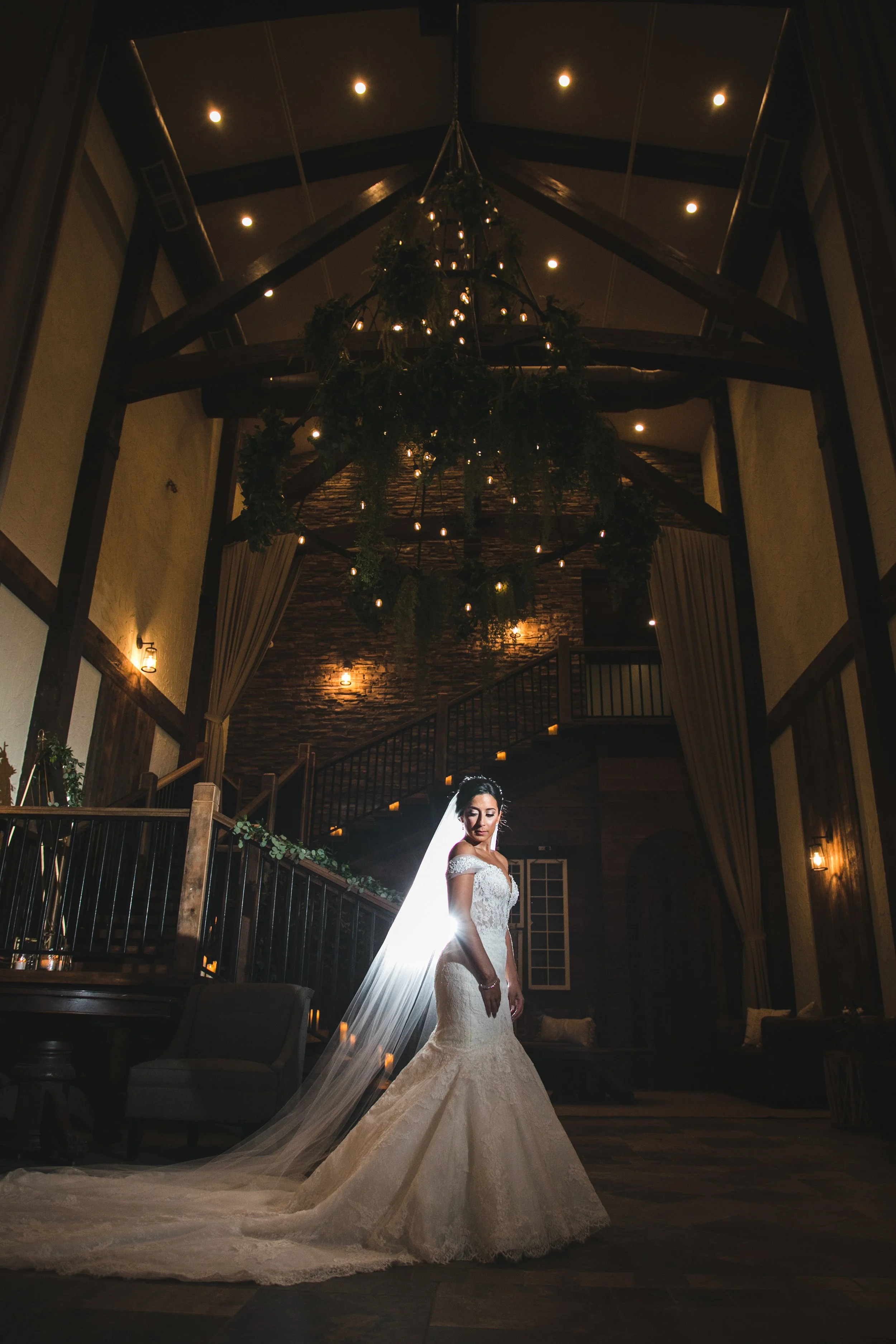 Bride standing underneath a big beautiful rustic circle chandelier that is covered in greenery in a rustic wedding venue