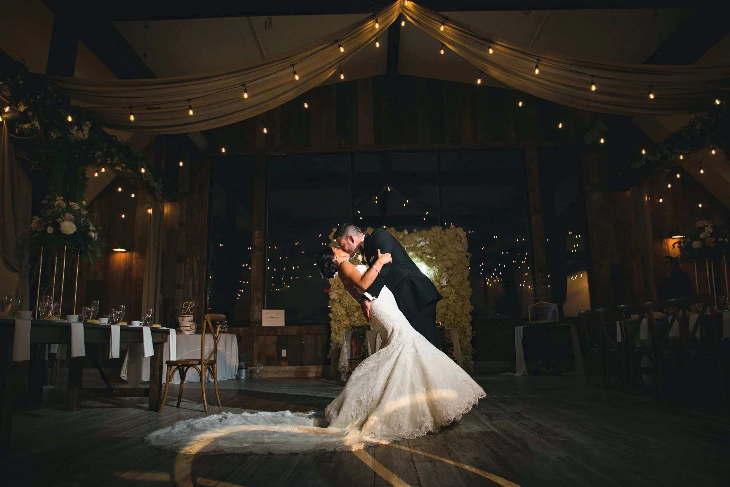 Groom kissing his bride while dipping her in front of their reception ballroom in their rustic barn wedding venue, with fairy lights overhead