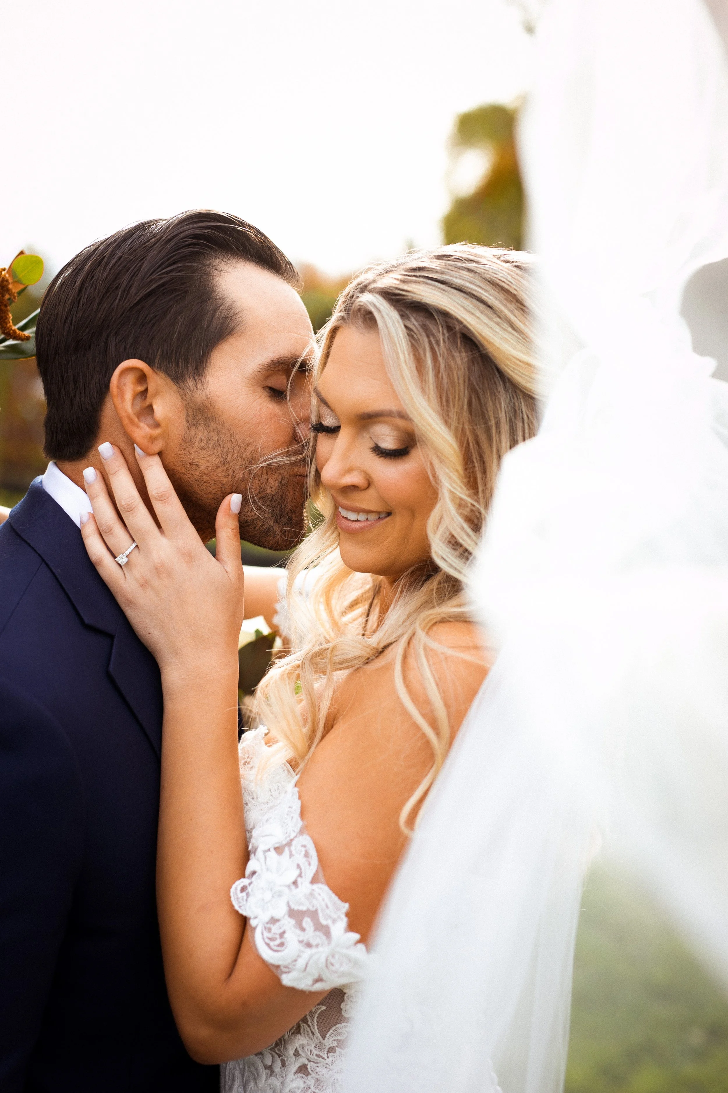 Close up of groom kissing bride on the cheek with her veil flowing toward the camera