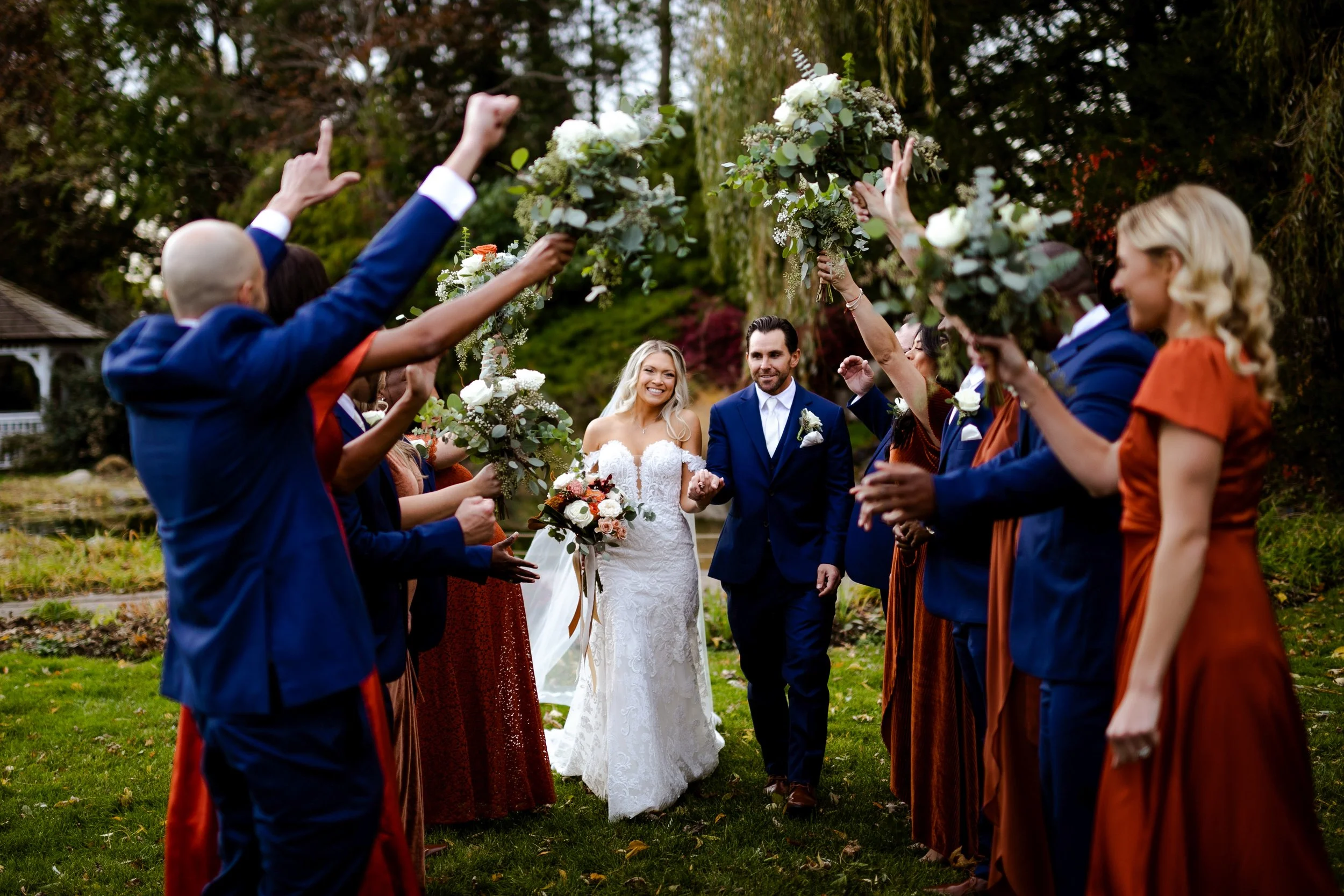 Bridesmaids wearing orange and red, and groomsmen wearing royal blue standing in two lines holding up their bouquets and hands while the bride and groom walk through between their two lineups