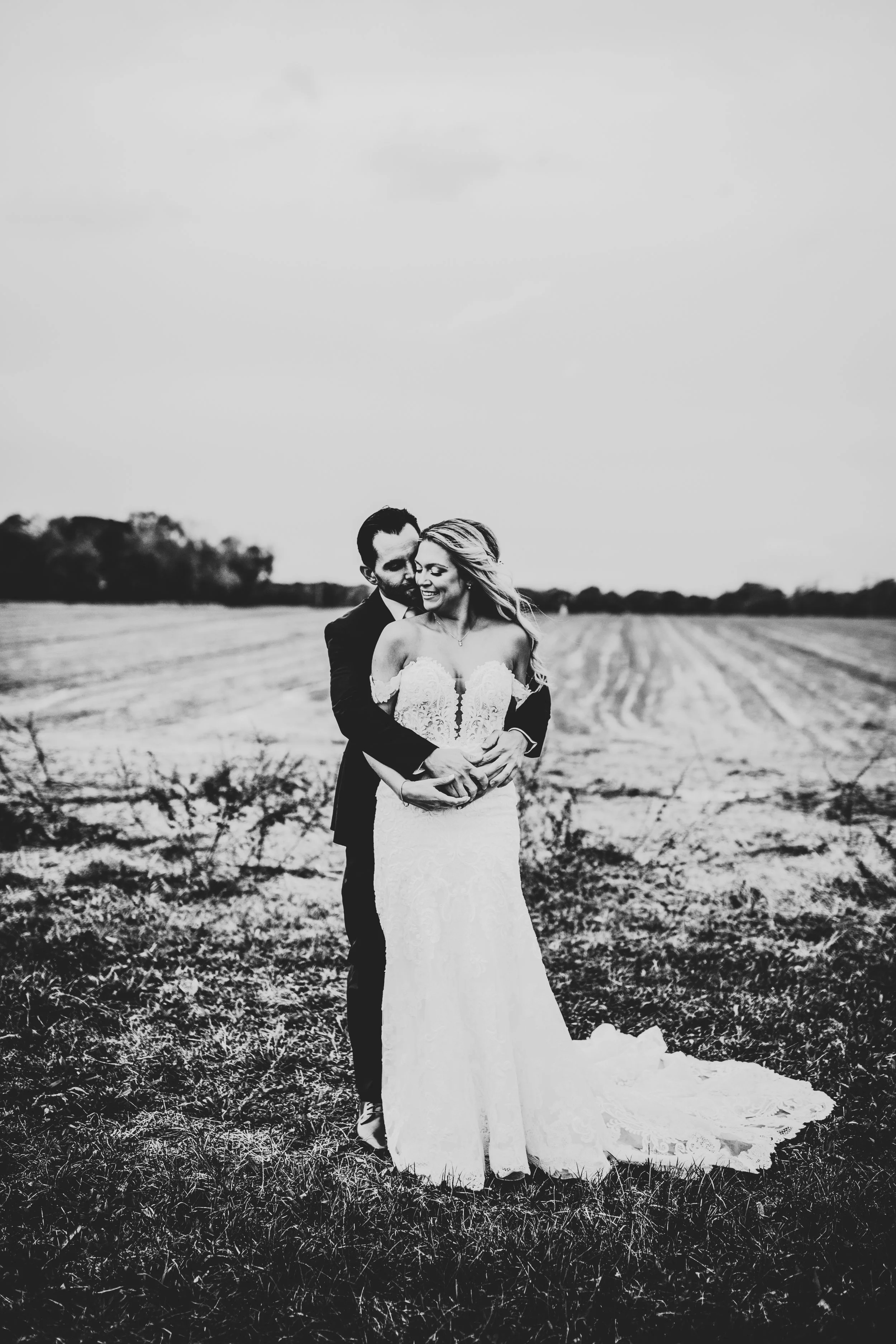 Groom hugging bride from behind wrapping arms around her in front of an empty field