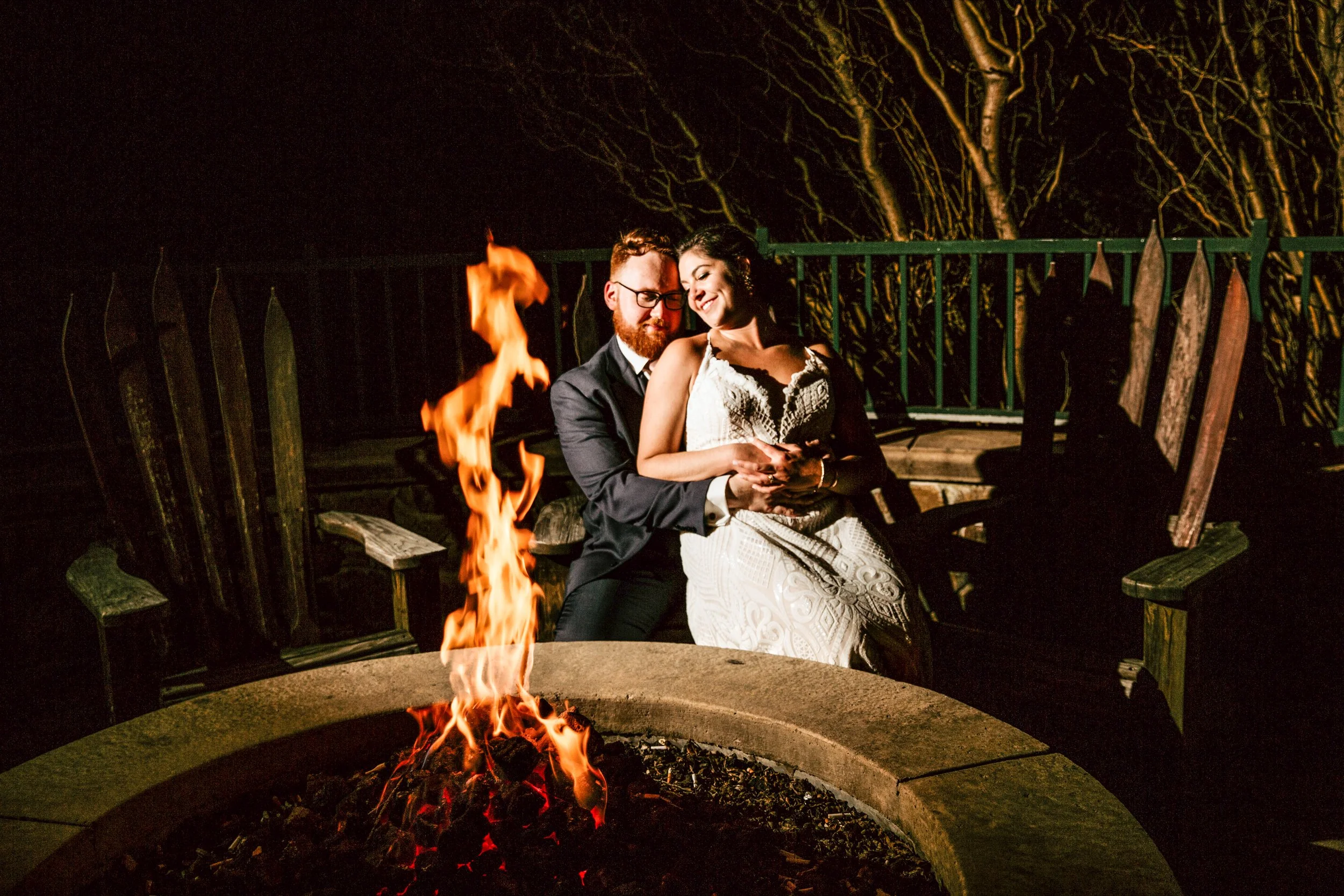 Bride and groom in an embrace sitting on a bench together outside in front of a flaming fire pit in the fall in New Jersey
