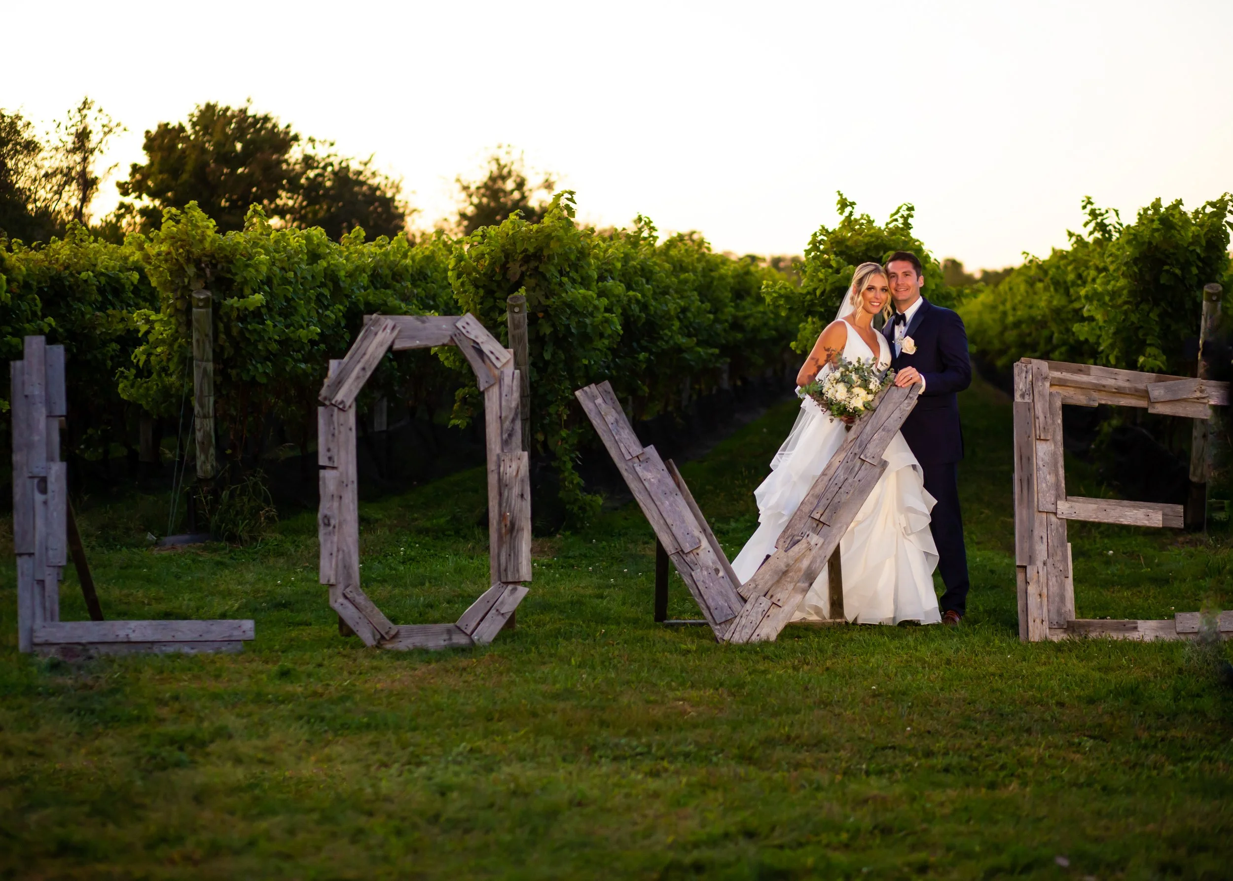 Bride and groom hugging and smiling behind a rustic wooden LOVE sign in front of a long island vineyard