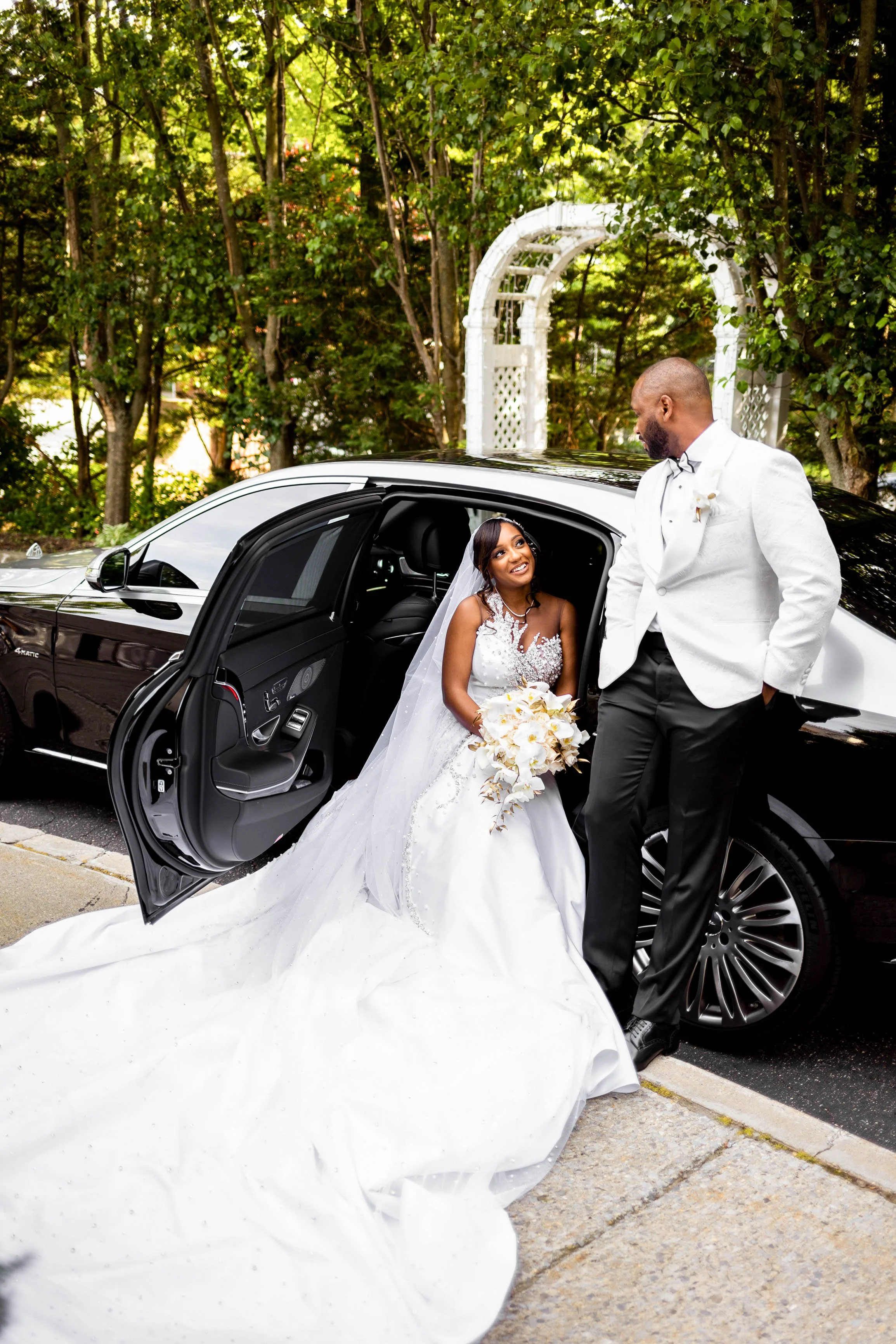 Gorgeous black couple wearing their wedding dress and tux, bride is sitting inside car, groom is standing outside looking down at her