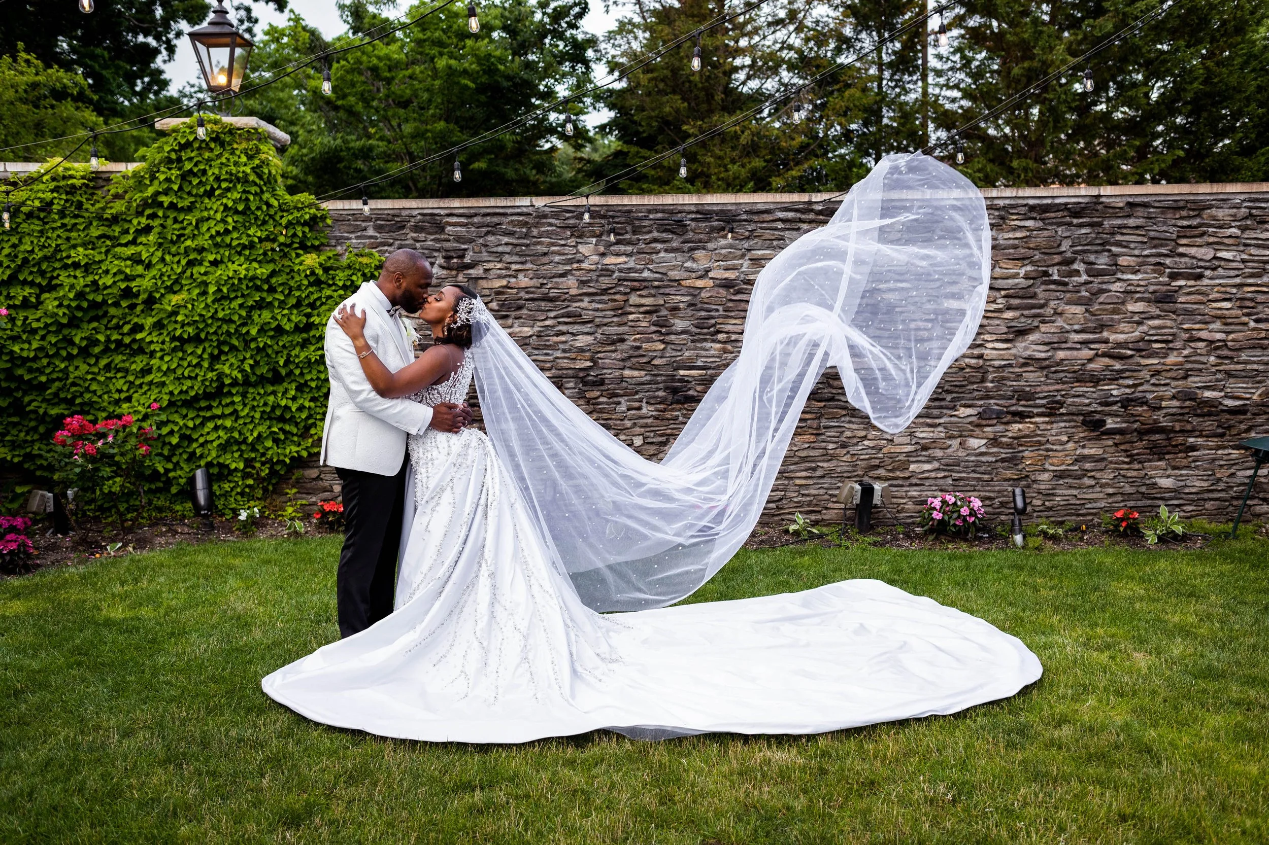 Bride and groom kissing in garden as bride’s dress and veil flow behind her