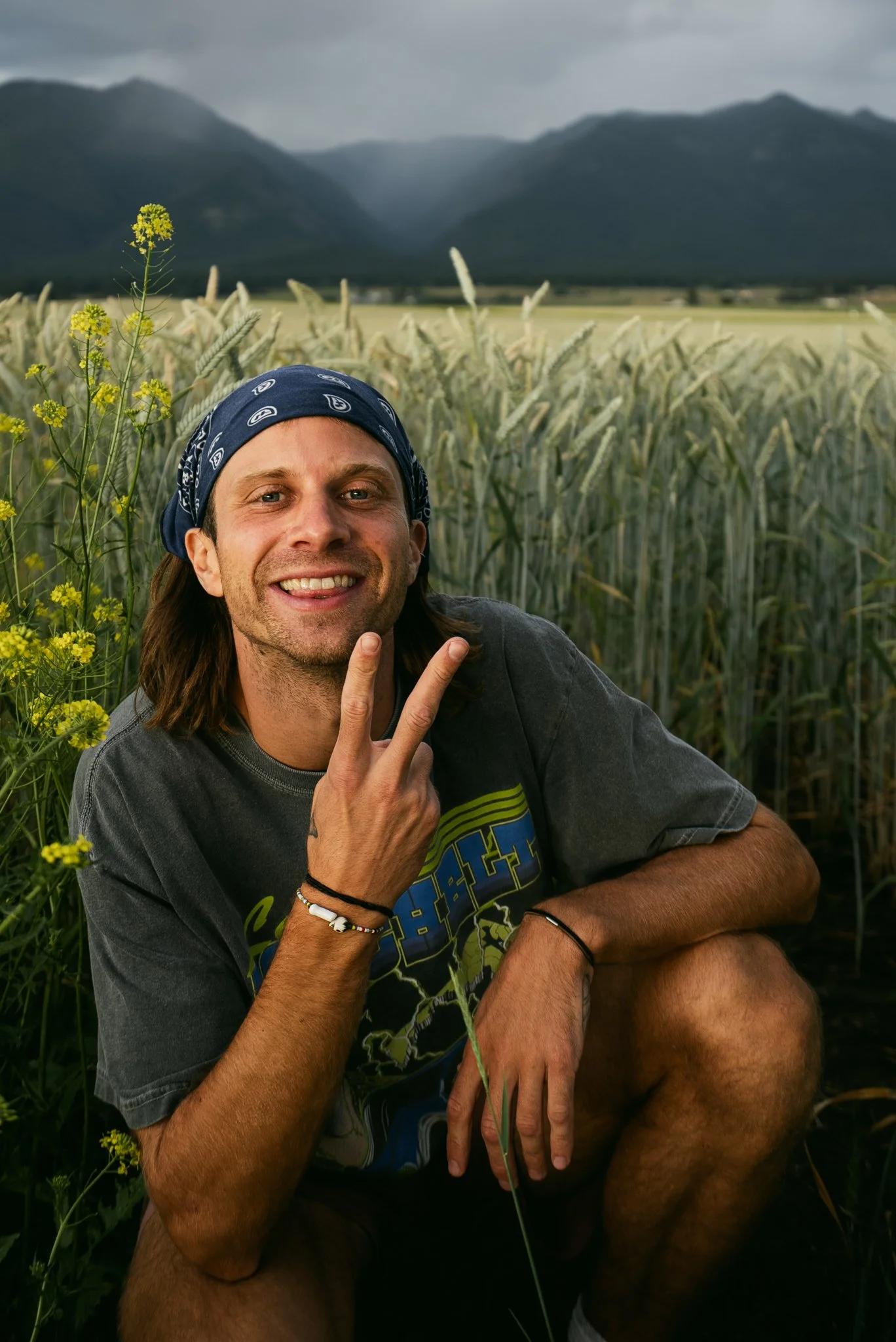 A smiling man with long hair and a bandana making a peace sign, crouching in a field of tall wheat with mountains in the background on a cloudy day.