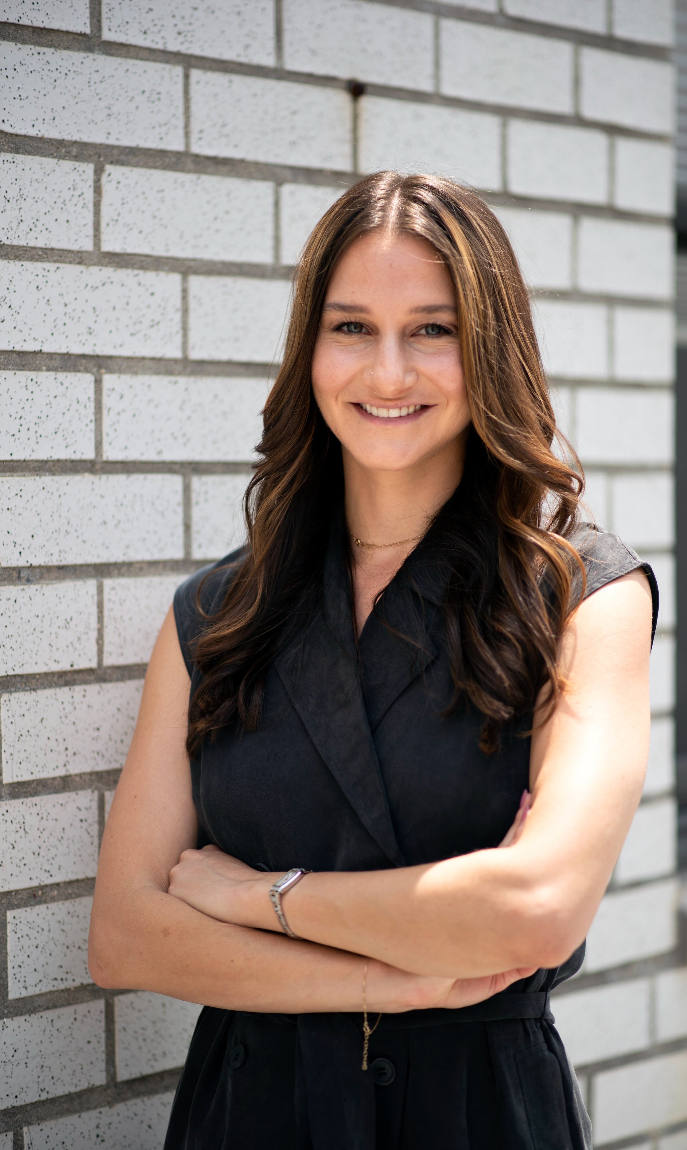 Therapist Alana wearing black with arms crossed in front of a white, brick wall.