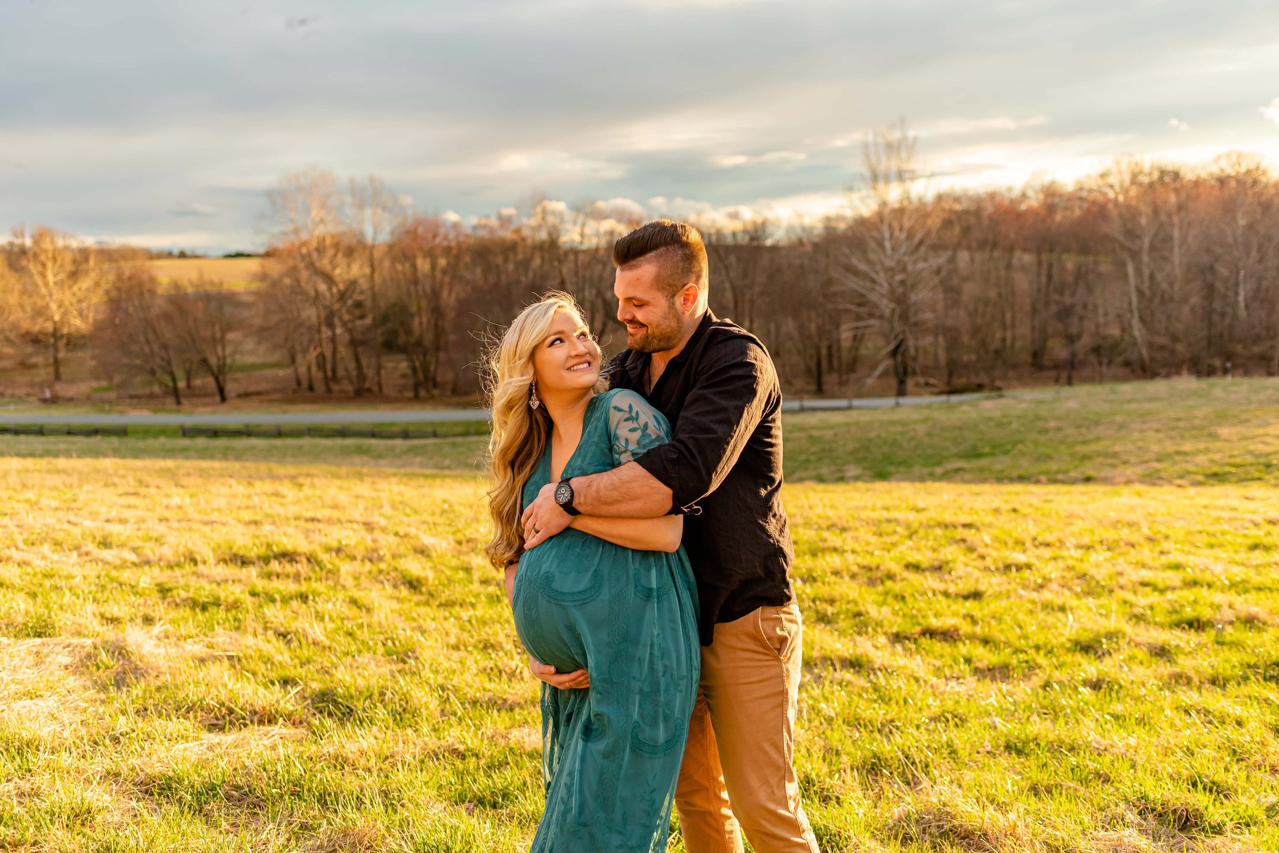 Maternity photos with couple standing in a field smiling at each other