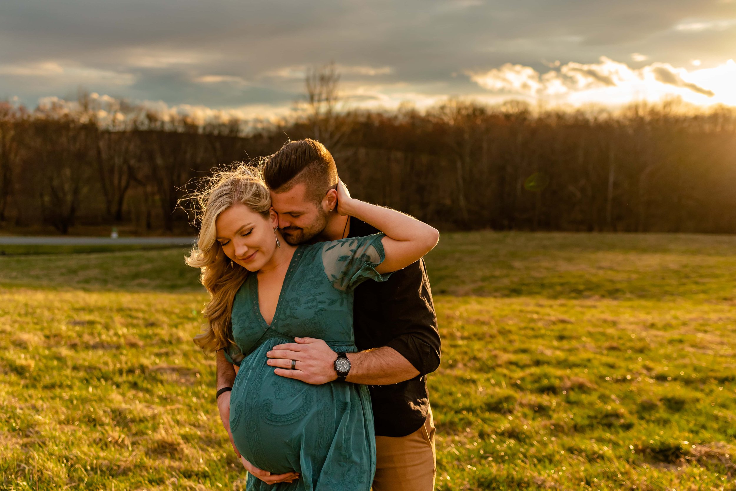 Maternity photos with couple standing in a field cuddling surrounded by sunset glow