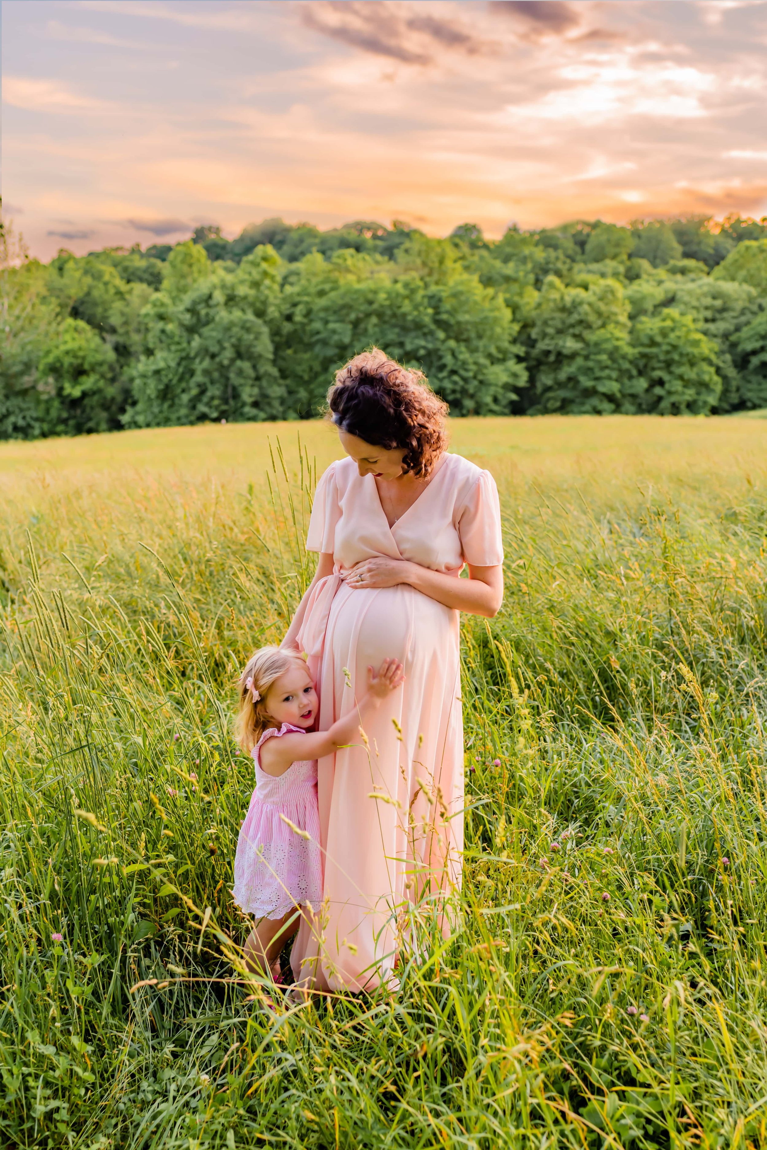 Maternity photo of pregnant woman standing in a field with older daughter holding onto her belly 