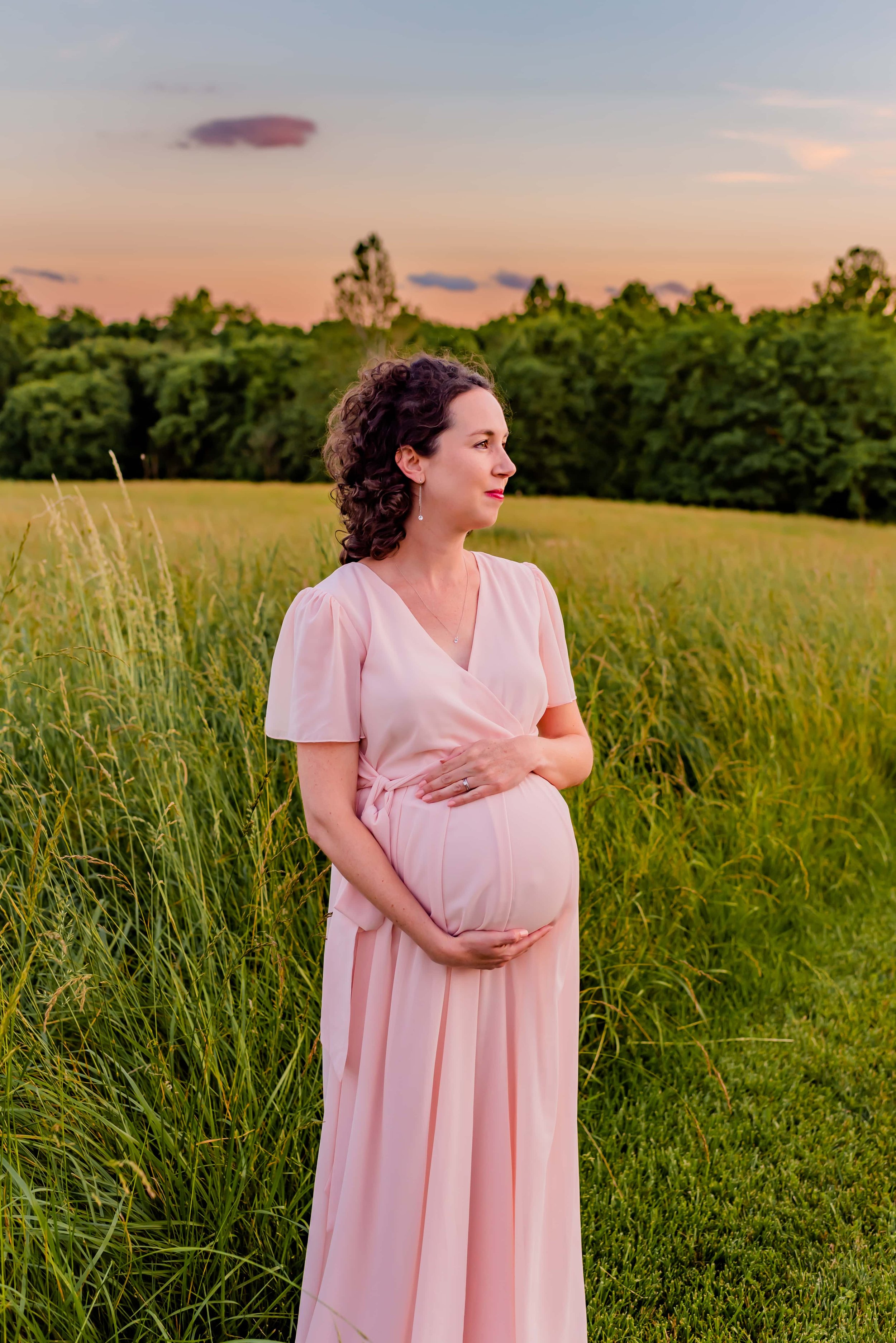 Maternity photo of woman standing in a field holding her belly and looking into the distance