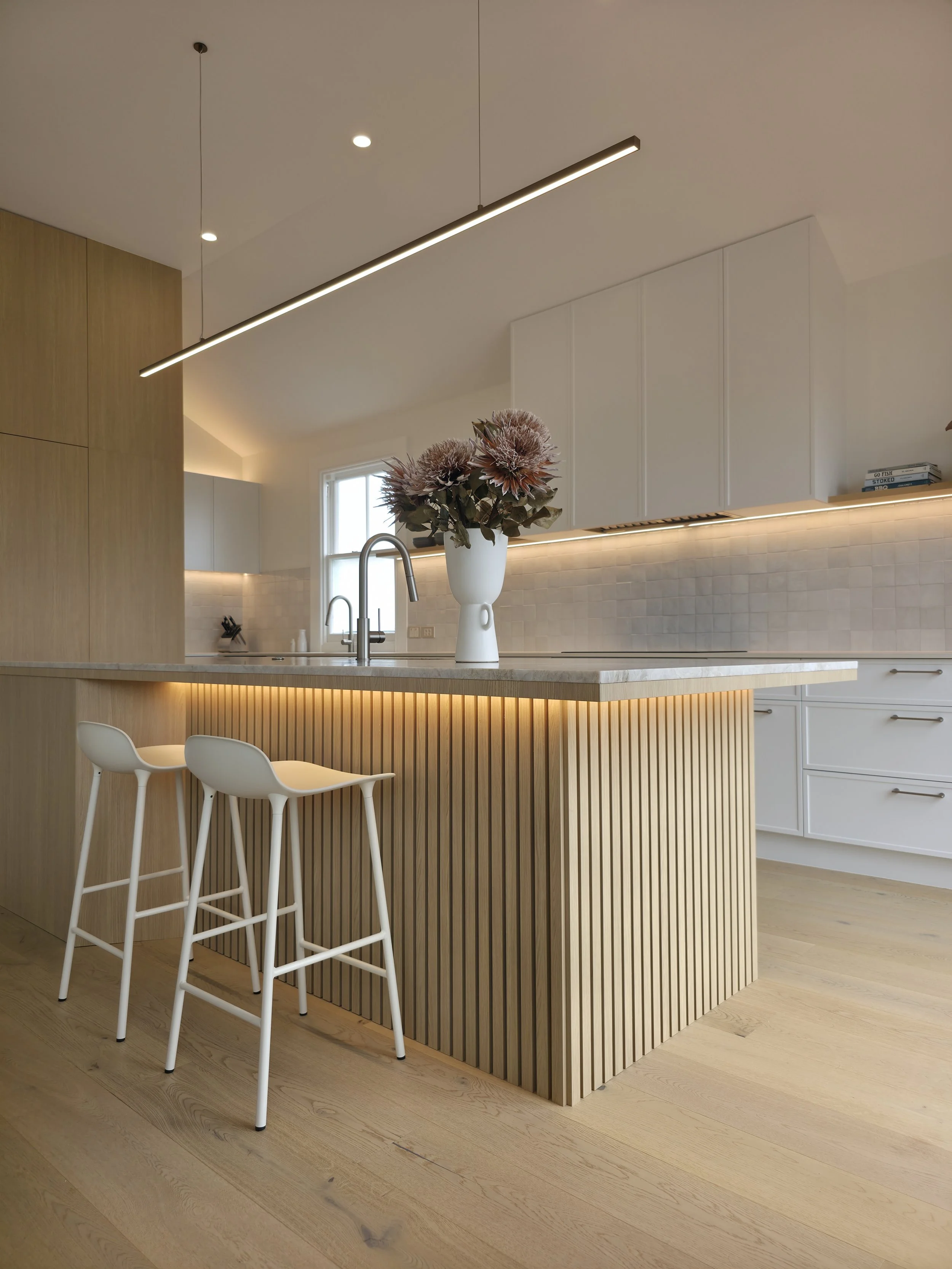 Modern kitchen with wooden and white cabinetry, a marble island with floral centerpiece, hanging linear light, and bar stools.