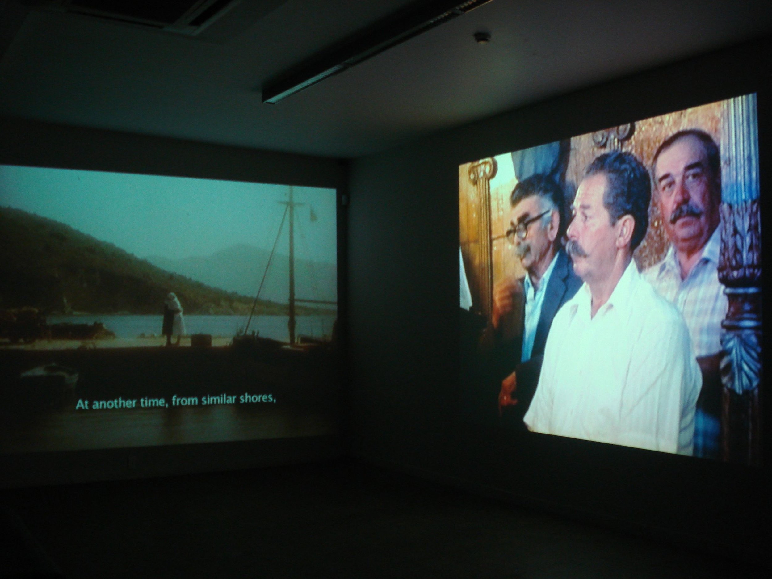 Projection of two side-by-side images on a wall. The left image shows a person standing by a waterfront with a boat mast and mountains in the background, featuring the subtitle "At another time, from similar shores." The right image displays three me