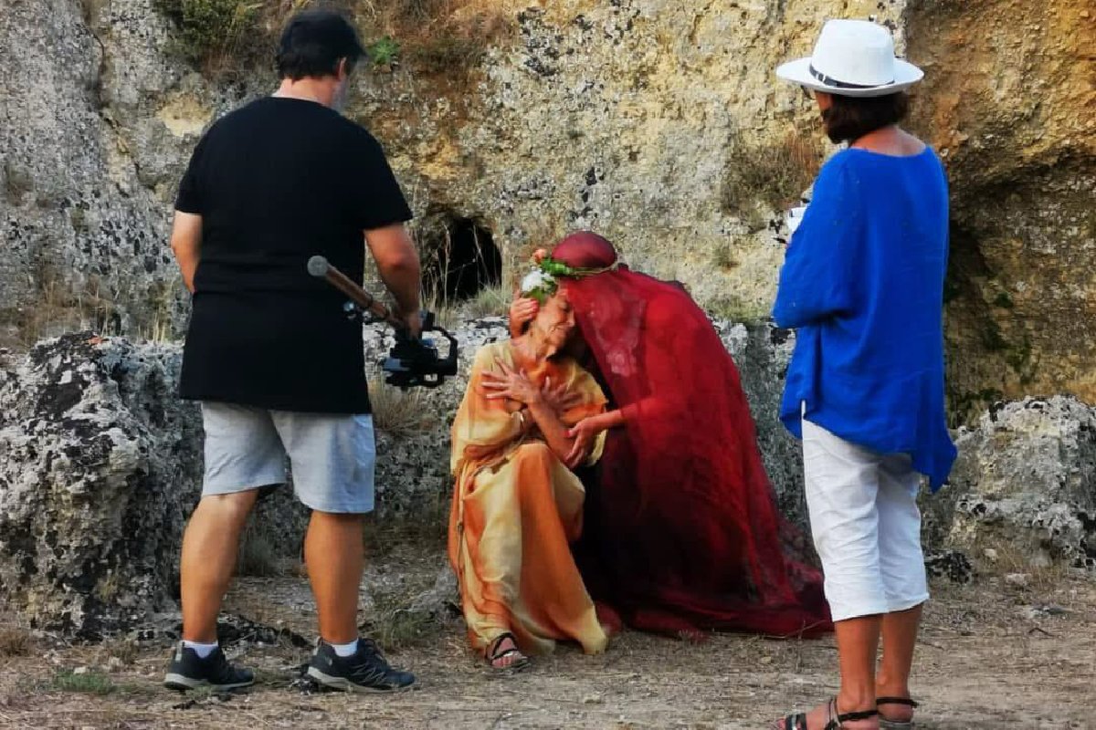 Outdoor film scene with crew, actors in historical costumes and floral crowns, and a camera operator.