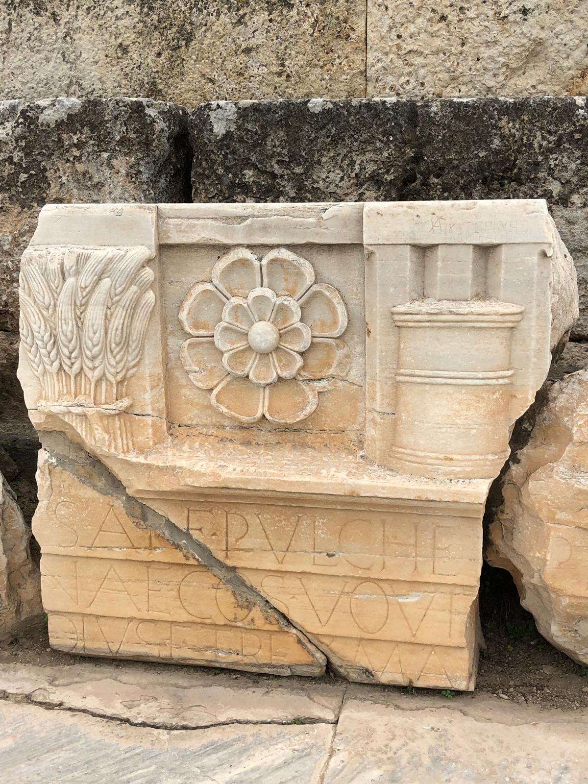 Ancient stone carving with decorative elements and Latin script, featuring flowers and wheat motif.